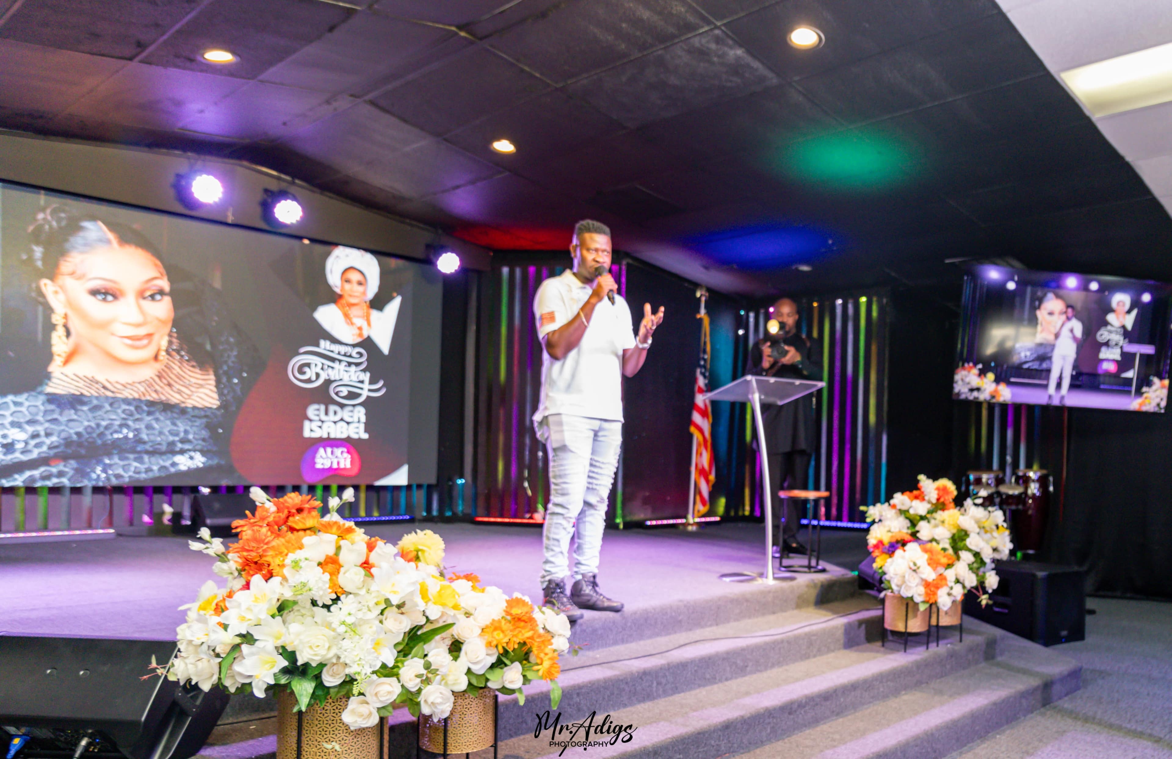 A speaker stands at a podium in a church setting, addressing an audience, with large screens displaying images and text in the background.