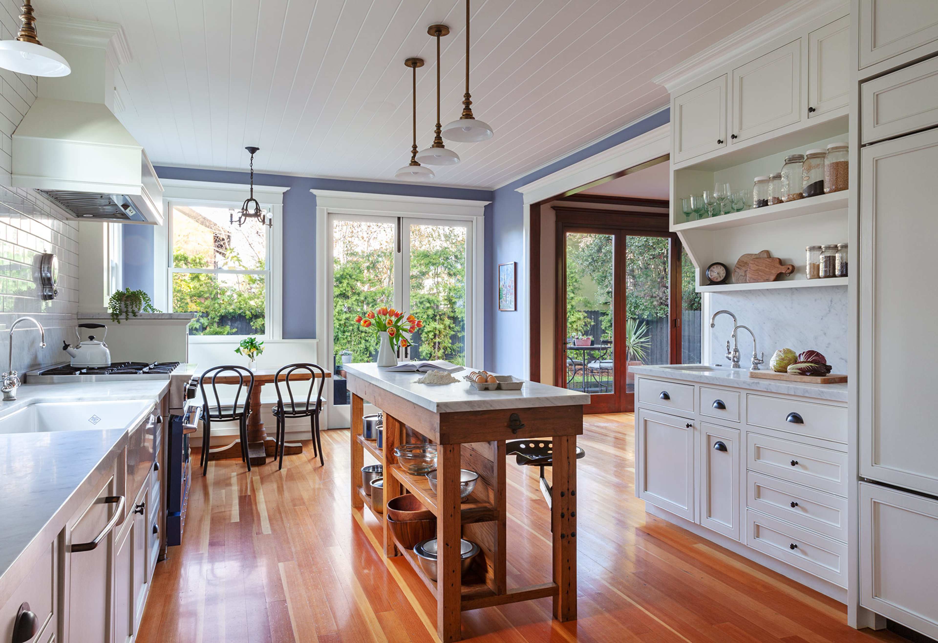 The kitchen features white cabinets, a large wooden island, and large windows overlooking greenery.
