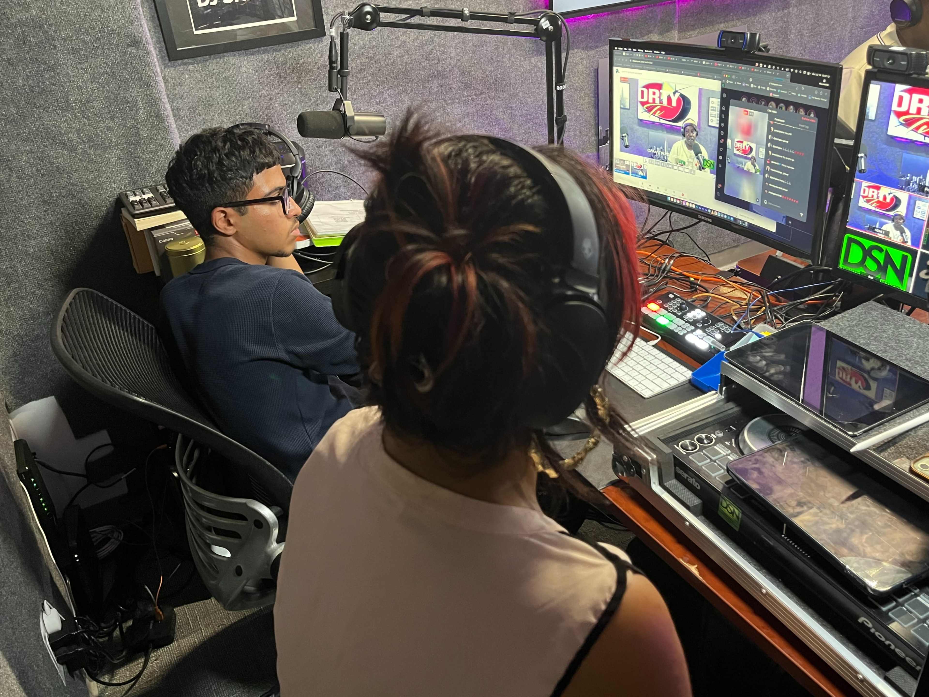 A young man and woman are seated in a recording studio, with the woman facing away from the camera and the man focused on a computer screen displaying a video.