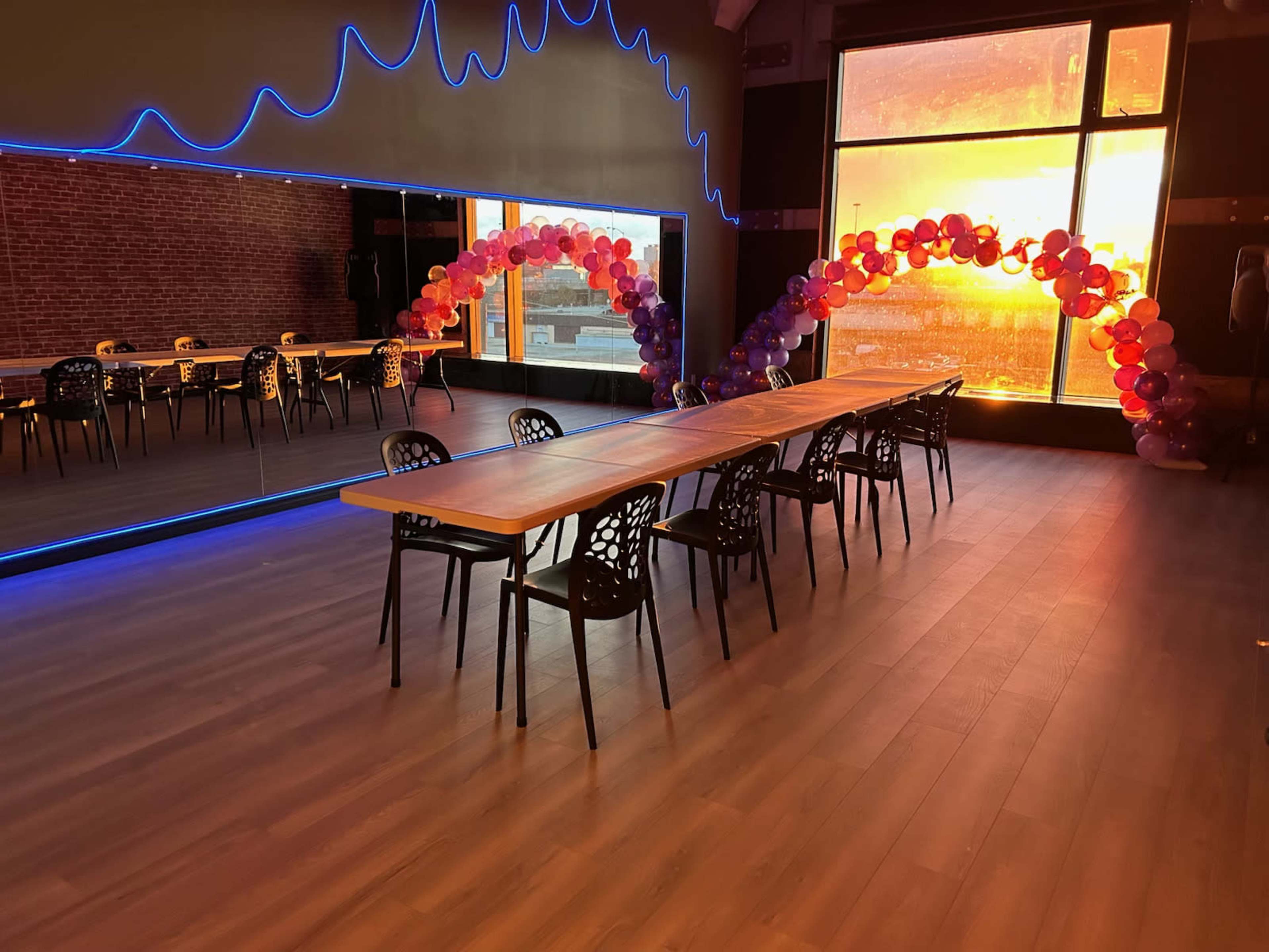 A spacious room features a long table surrounded by chairs, with a backdrop of colorful balloon arches and a sunset visible through large windows.