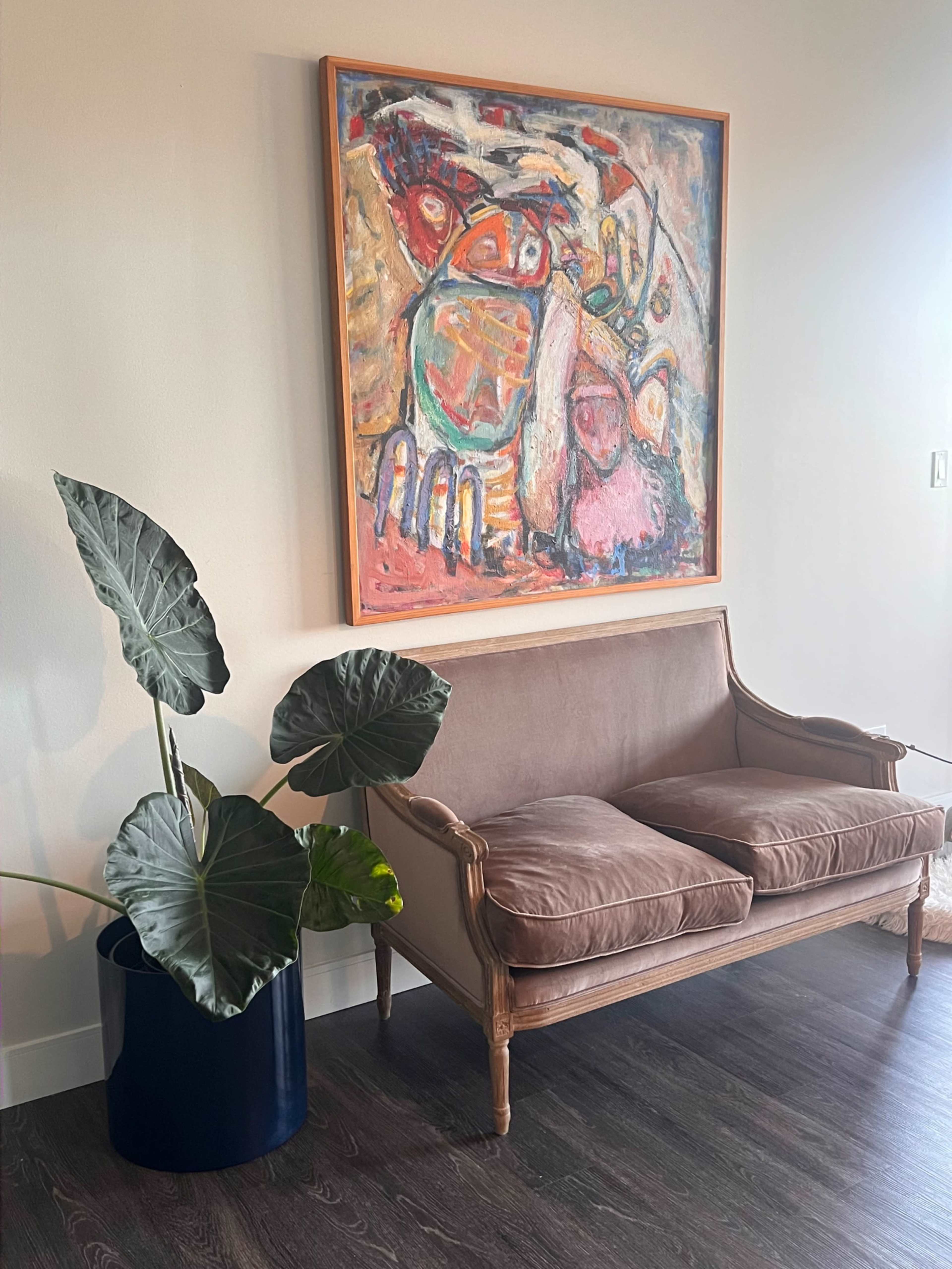 A brown upholstered loveseat sits beside a large green plant in a blue pot, under a colorful abstract painting on the wall.