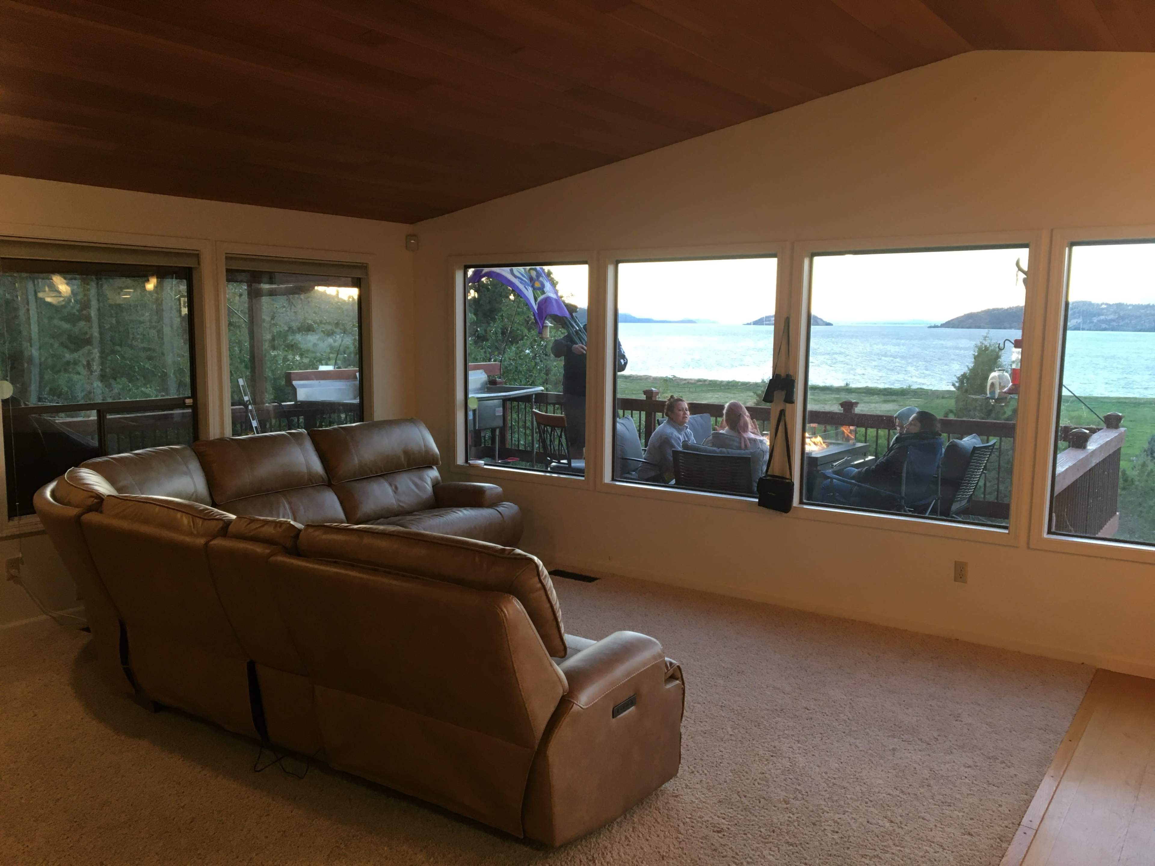 A living room featuring a leather couch faces large windows that overlook a patio with people gathered by a fire pit beside a body of water.
