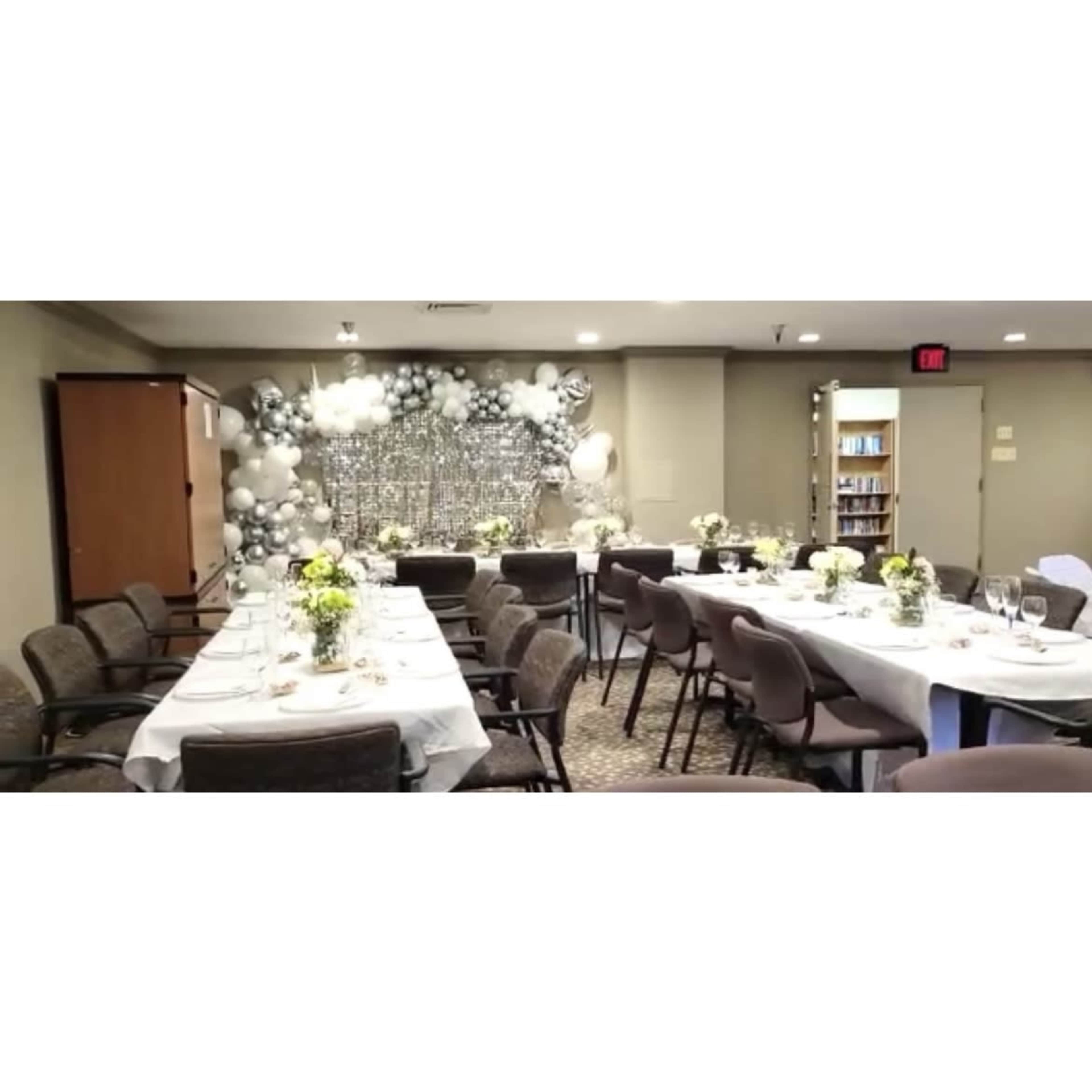 The image shows a banquet room set up for an event, featuring long tables covered with white tablecloths and decorated with floral centerpieces, with a backdrop of balloons and silver decor.