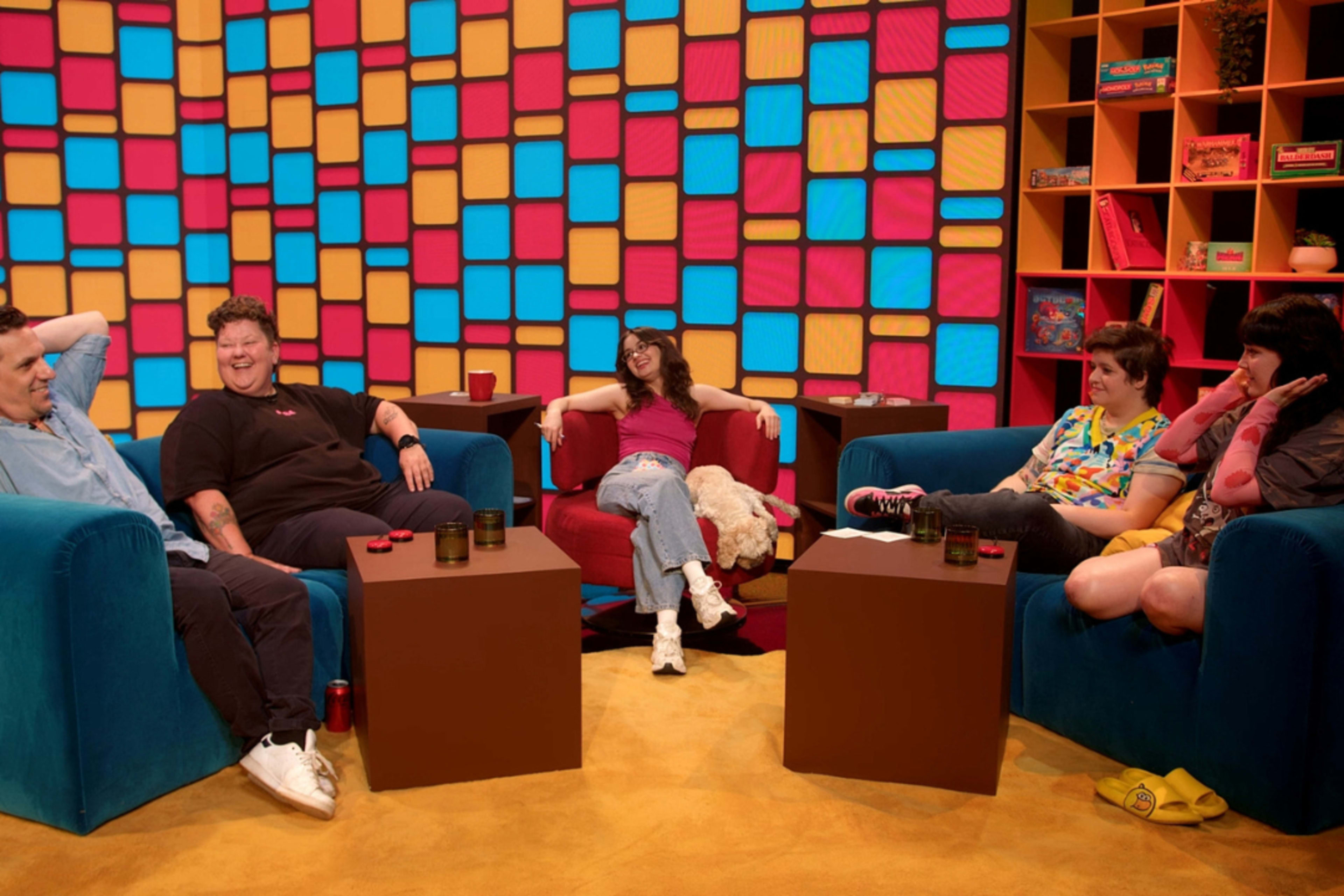 A group of five people sits in a colorful studio setting with patterned walls and modern furniture, engaging in conversation.
