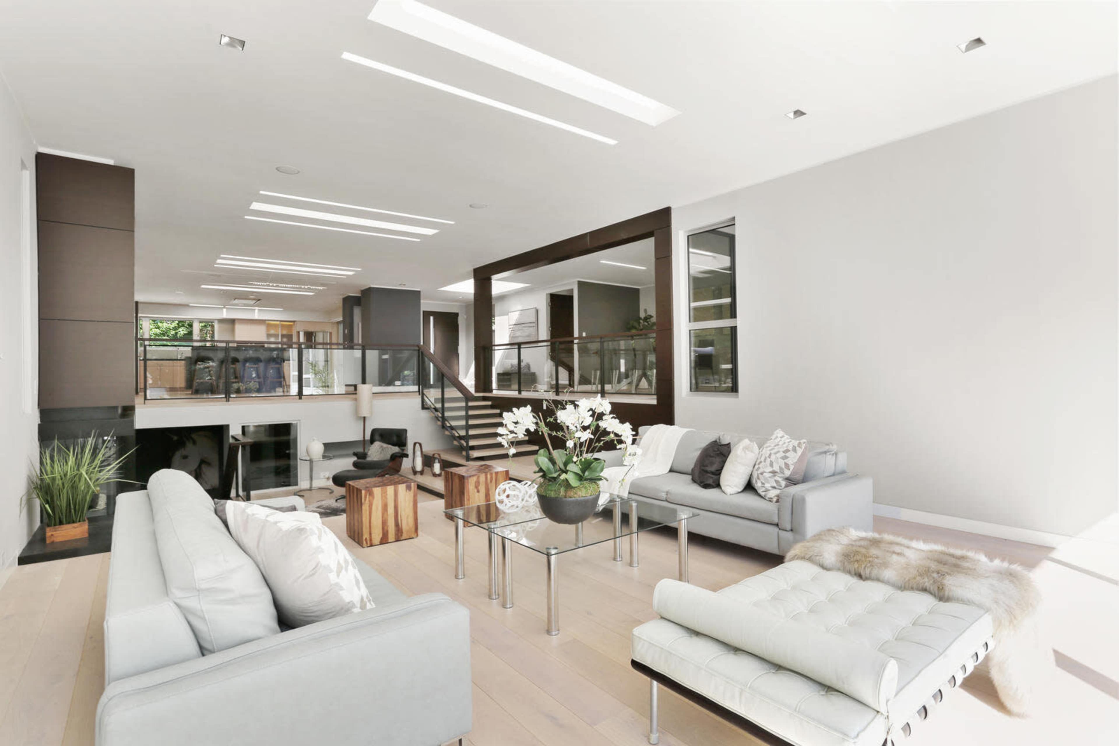 The scene shows a modern living room featuring gray sofas, a glass coffee table with a plant arrangement, and large windows that allow natural light to fill the space.