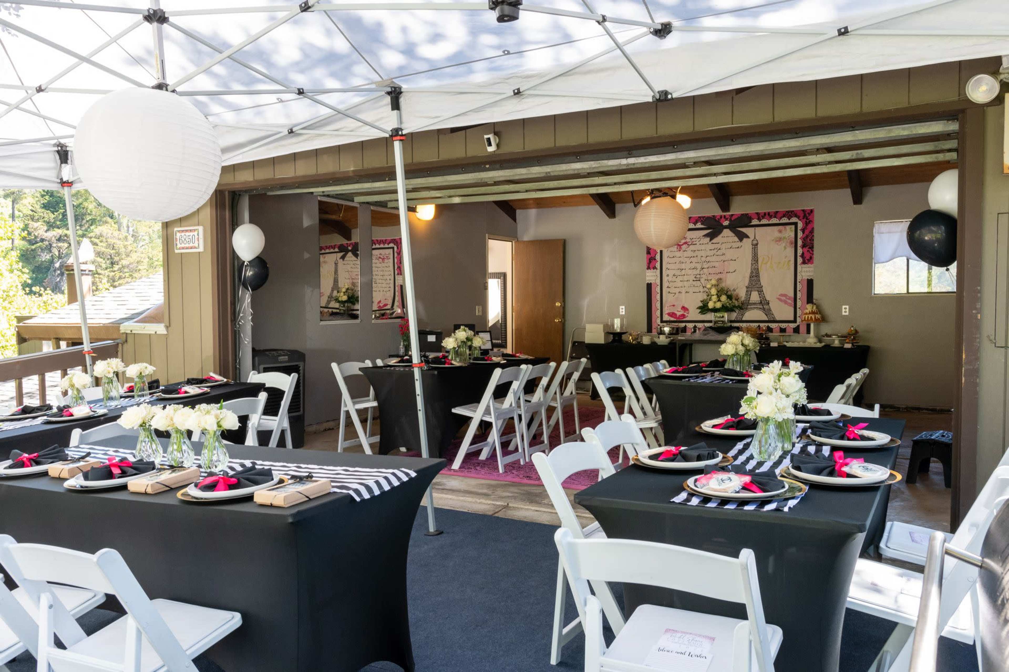 The image shows a decorated outdoor event space with black tablecloths, white chairs, and floral centerpieces under a tent.