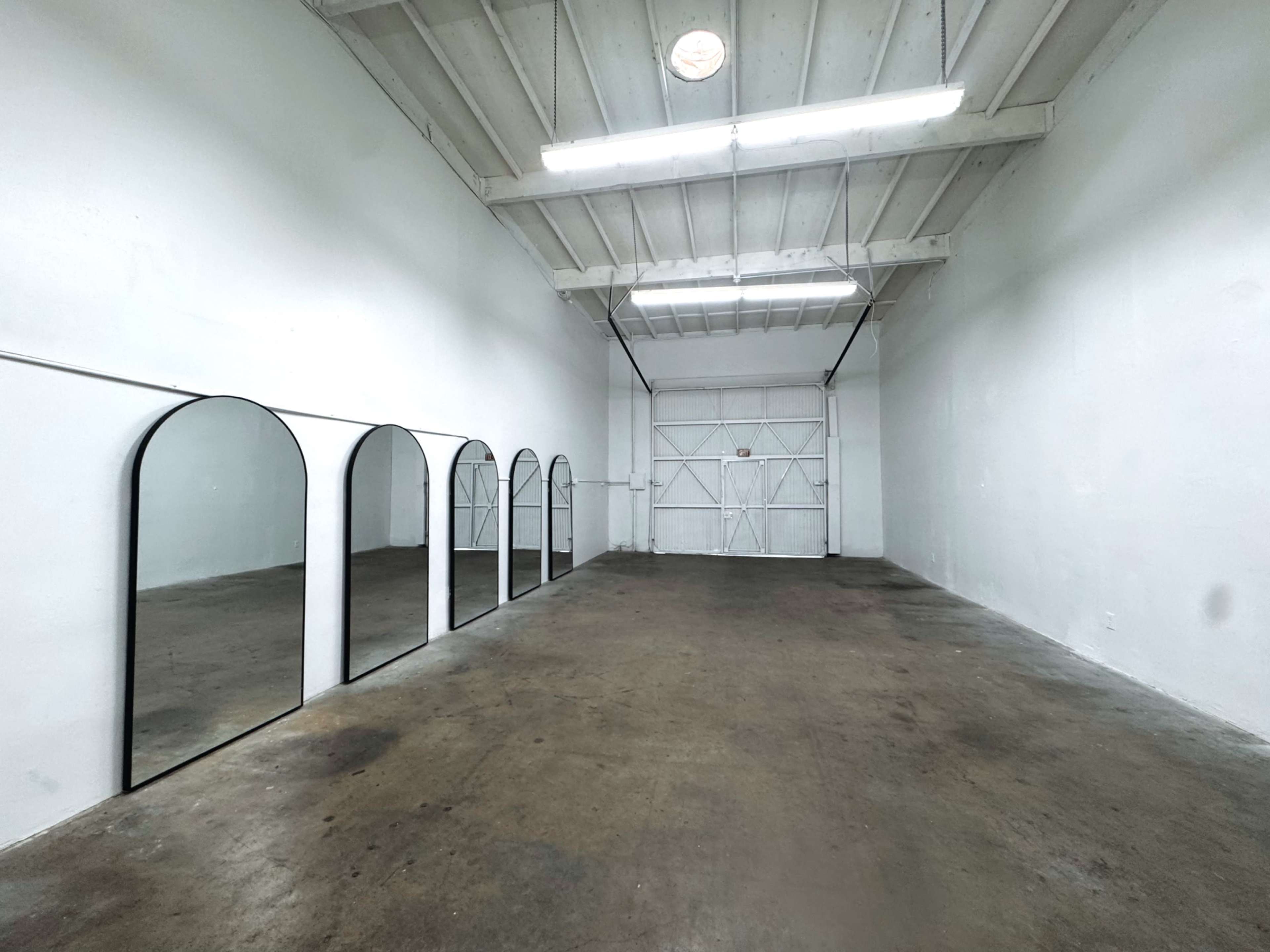 The image shows a large, empty room with white walls and arched mirrors mounted on one side, illuminated by overhead lights.