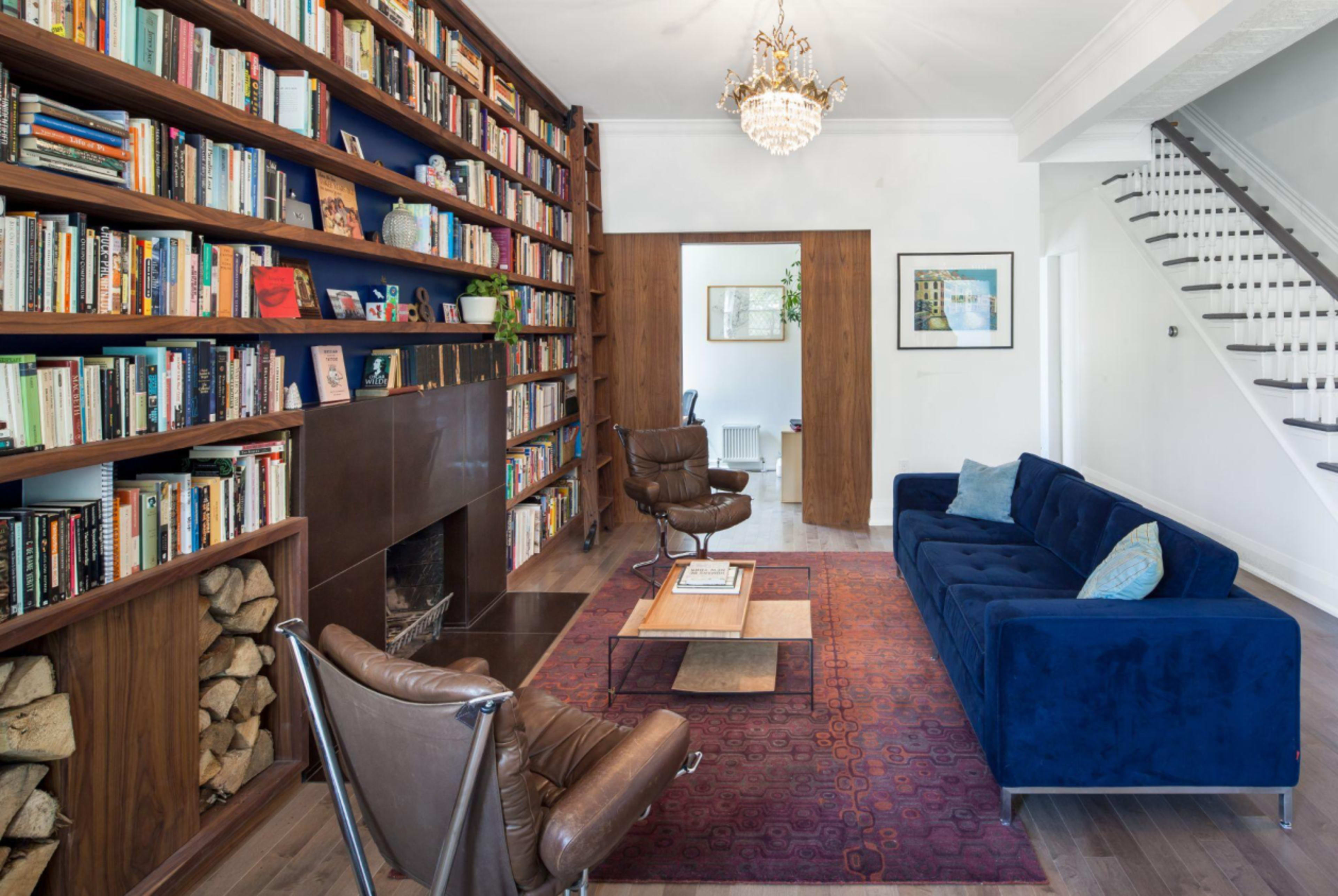 A modern living room features a blue couch, bookshelves filled with books, a wooden coffee table, and a chandelier above, with a staircase visible in the background.