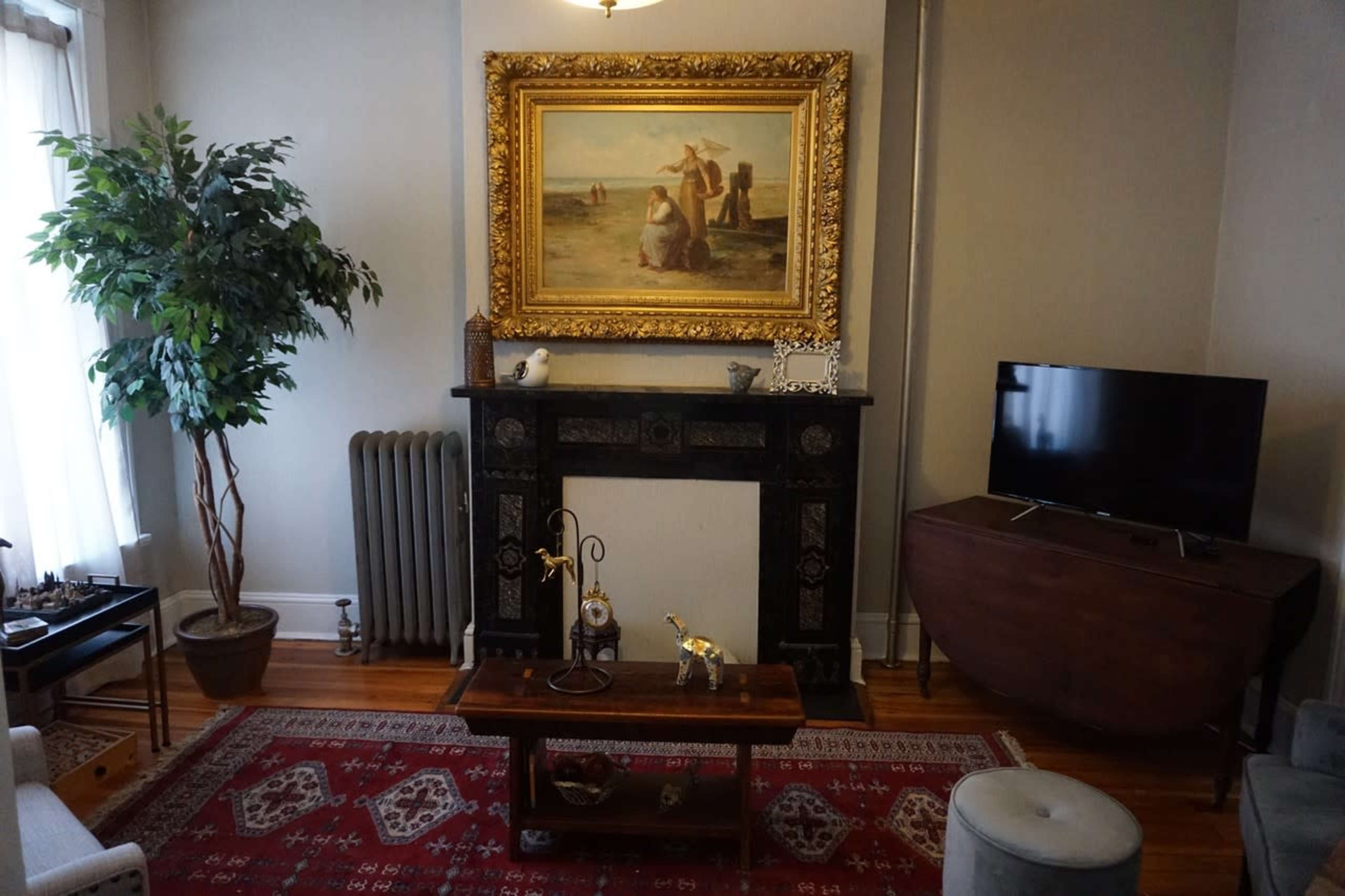 A living room features a large ornate painting above a black mantel, with a plant, a vintage radiator, a television, and a wooden coffee table on a red rug.