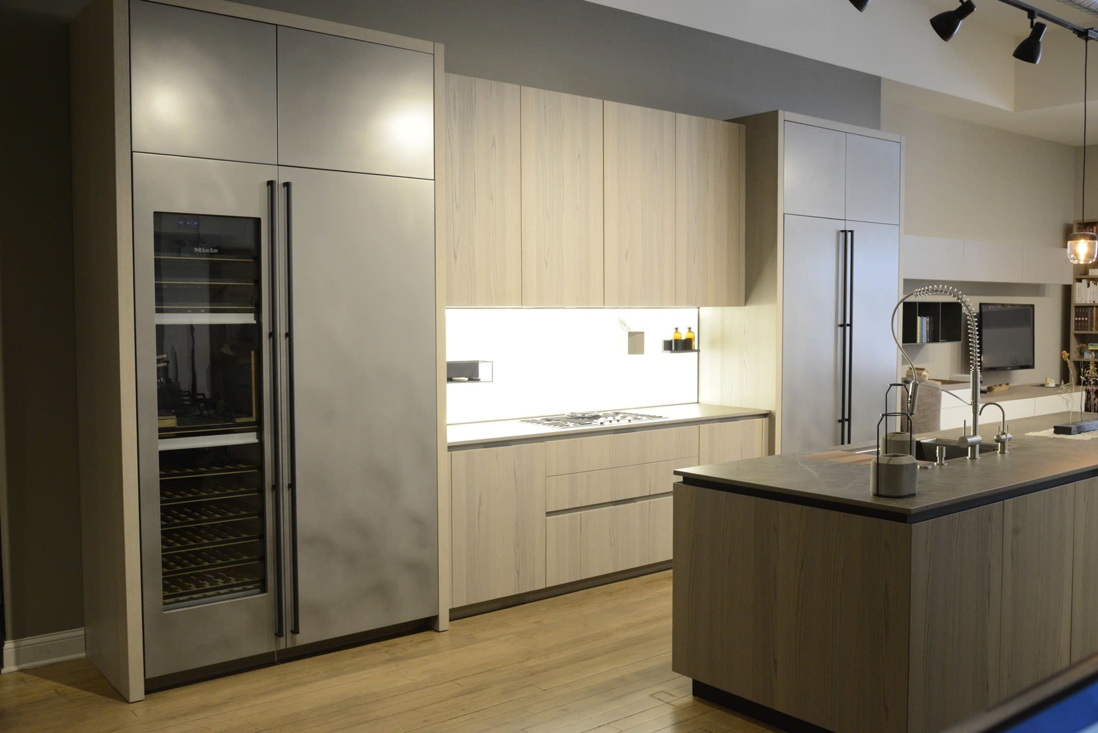 The image shows a modern kitchen with wooden cabinetry, a large stainless steel refrigerator, and an island with a sink.