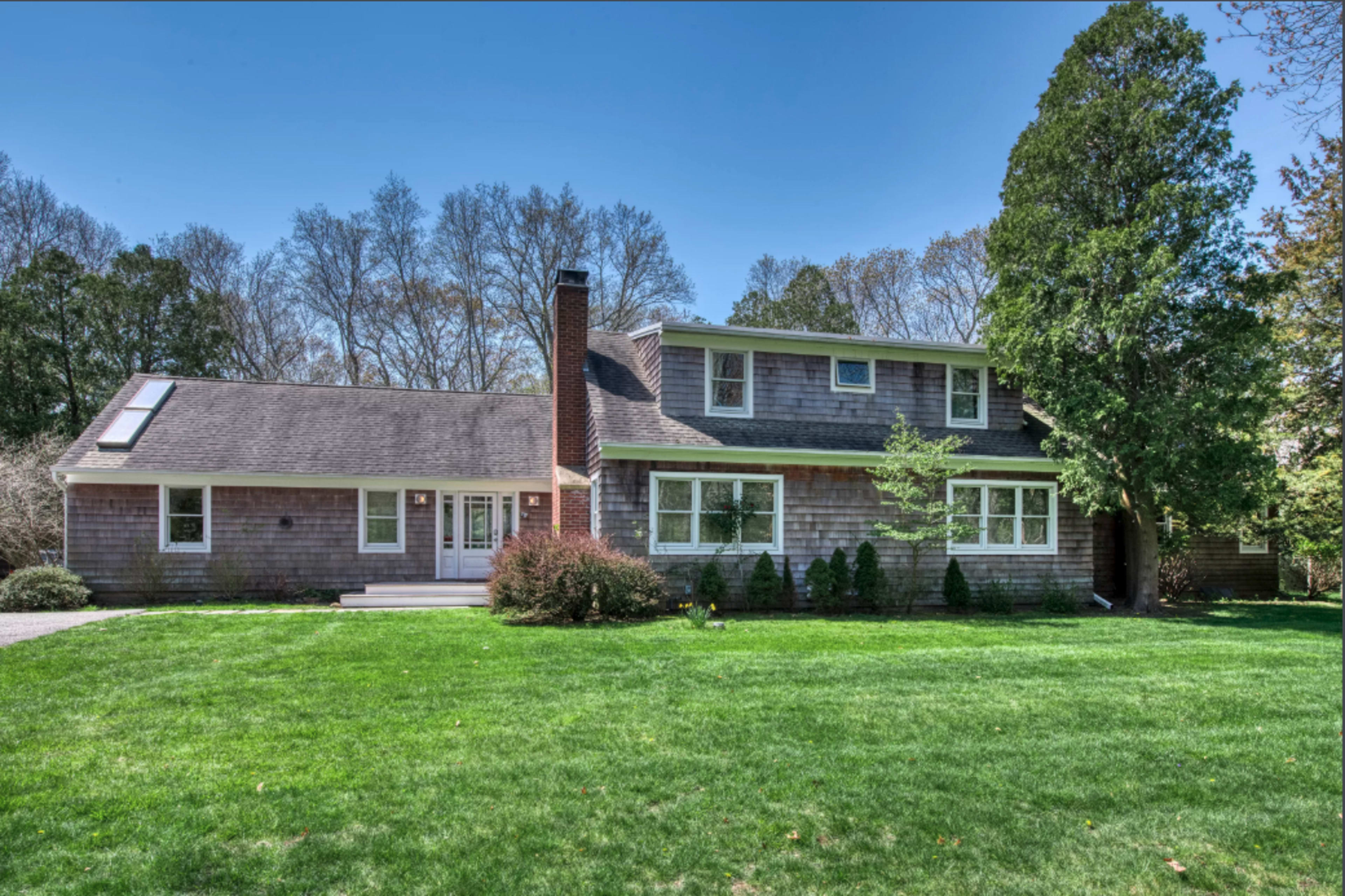 A two-story house with a shingle exterior and a large front lawn is surrounded by trees.