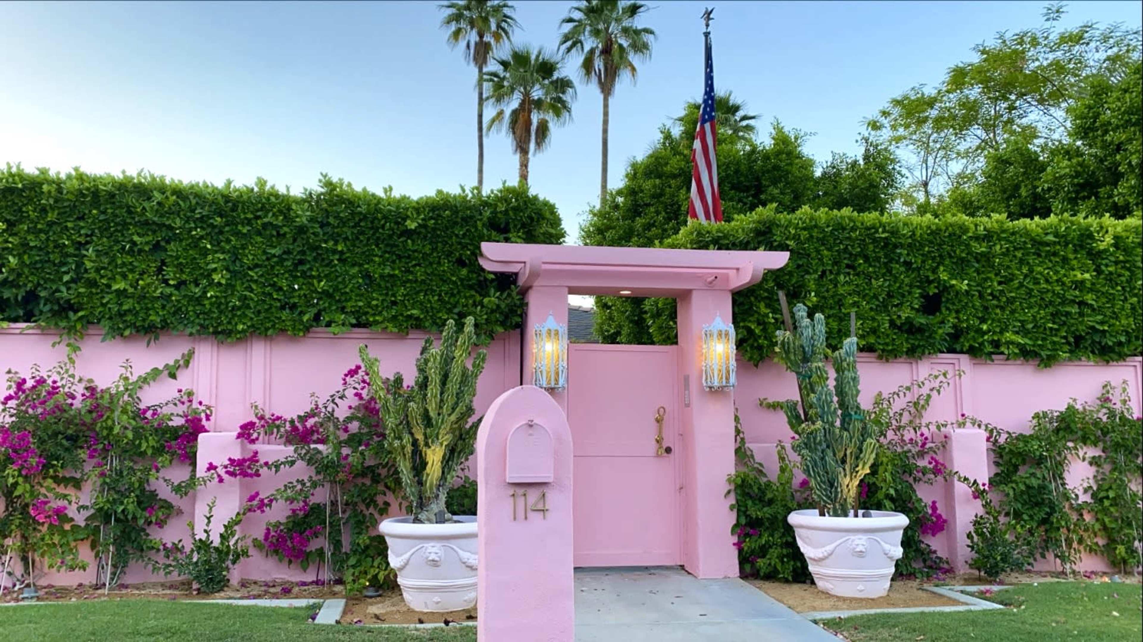 A pink entrance gate adorned with lanterns and surrounded by lush greenery and flowering plants features the number 114 and an American flag in the background.