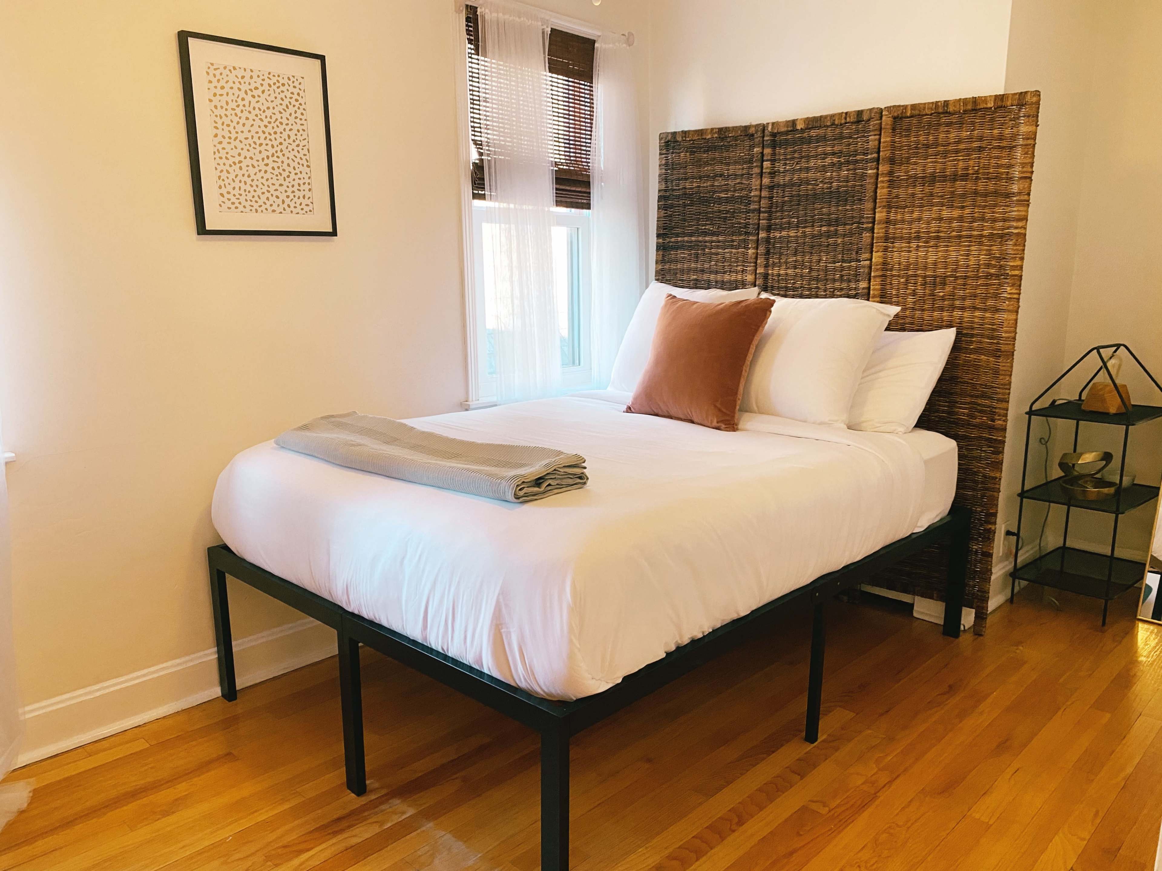 A minimalist bedroom features a bed with a woven headboard, white bedding, and a folded gray blanket, complemented by a framed artwork on the wall and a small decorative shelf.