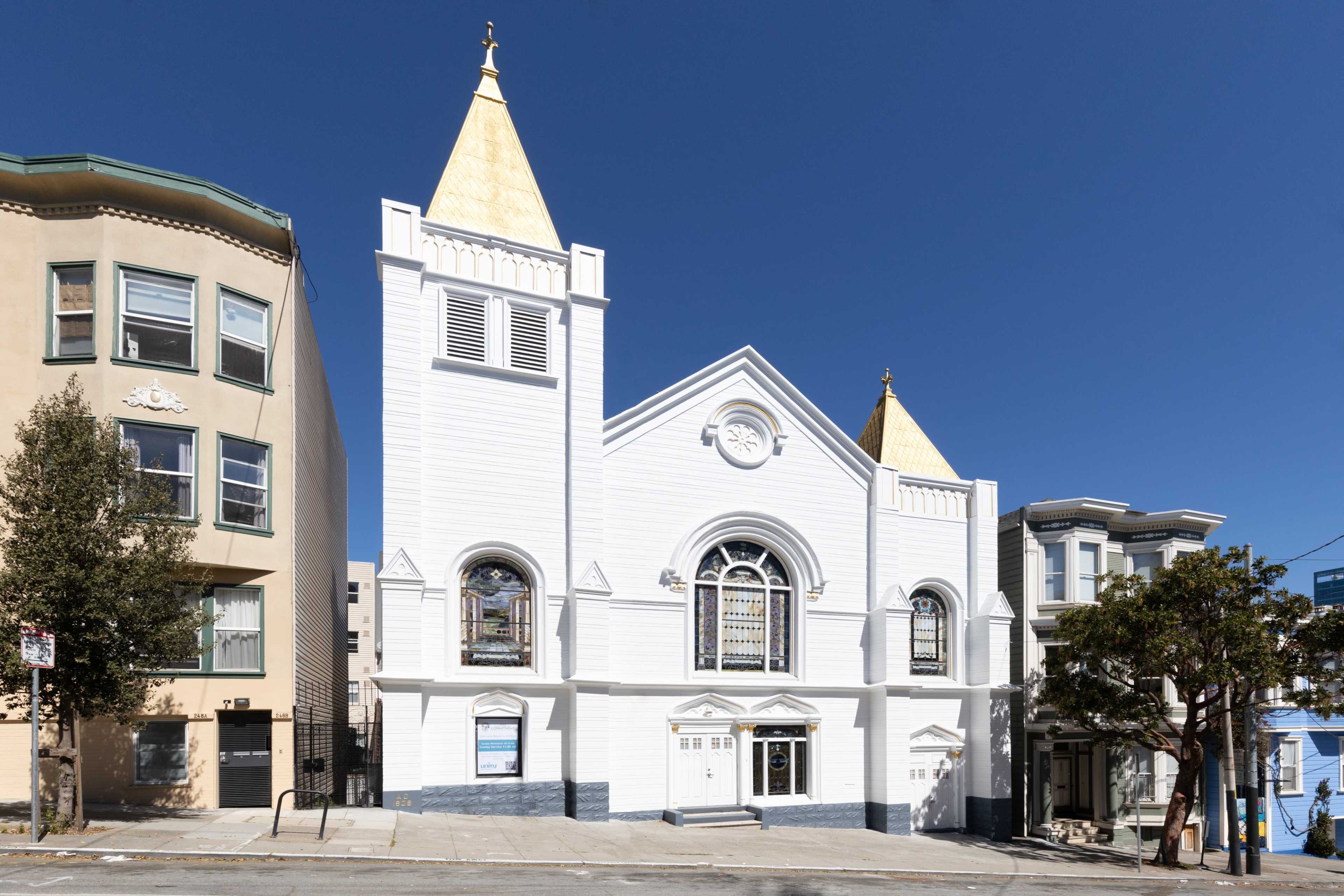 A white church with two golden spires situated at the corner of a street, flanked by two residential buildings.