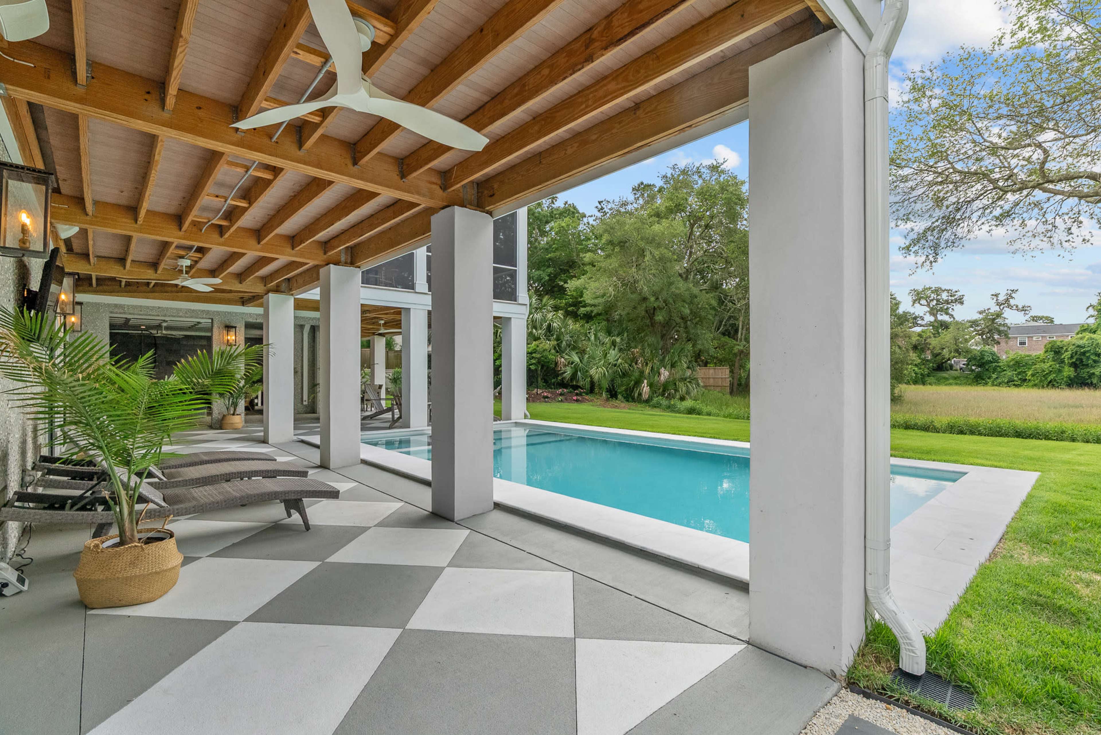 The image shows a spacious outdoor area featuring a swimming pool, paved with a checkered pattern, surrounded by lush greenery and tall trees.