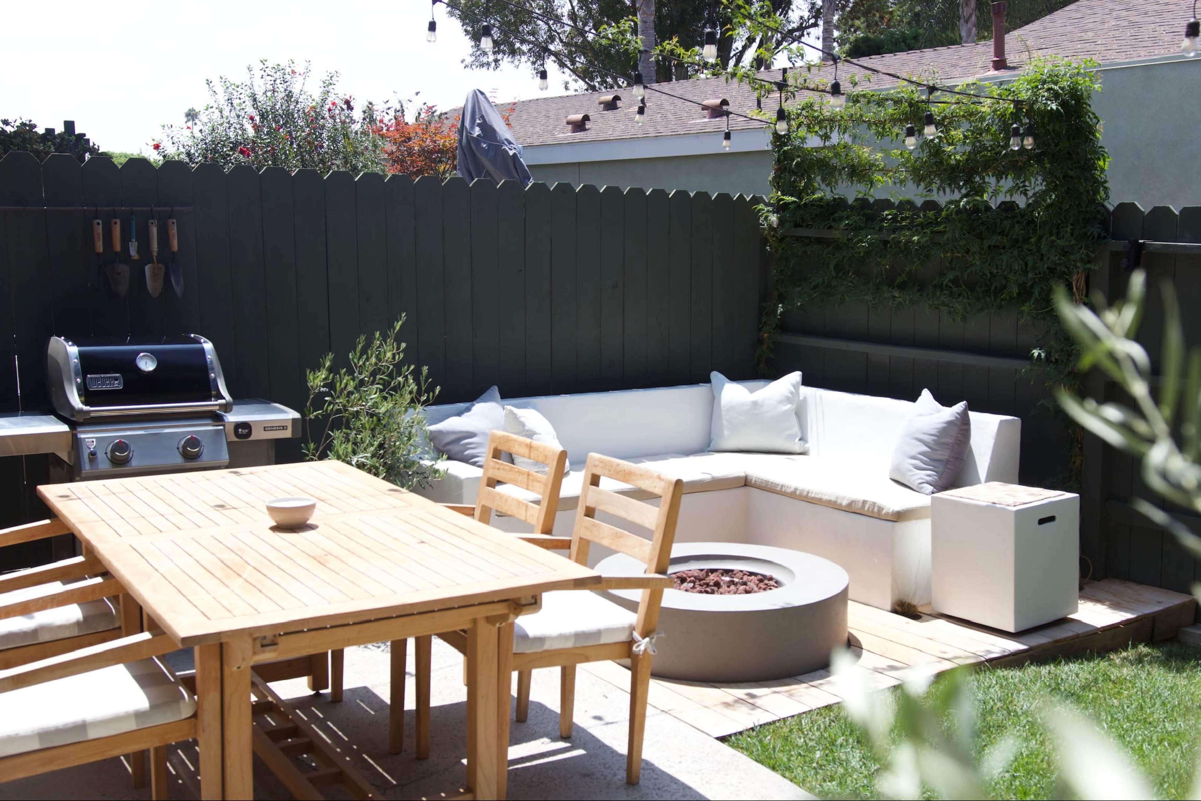 The image shows a backyard patio with a wooden dining table, a sectional sofa, a fire pit, and a grill surrounded by lush greenery.