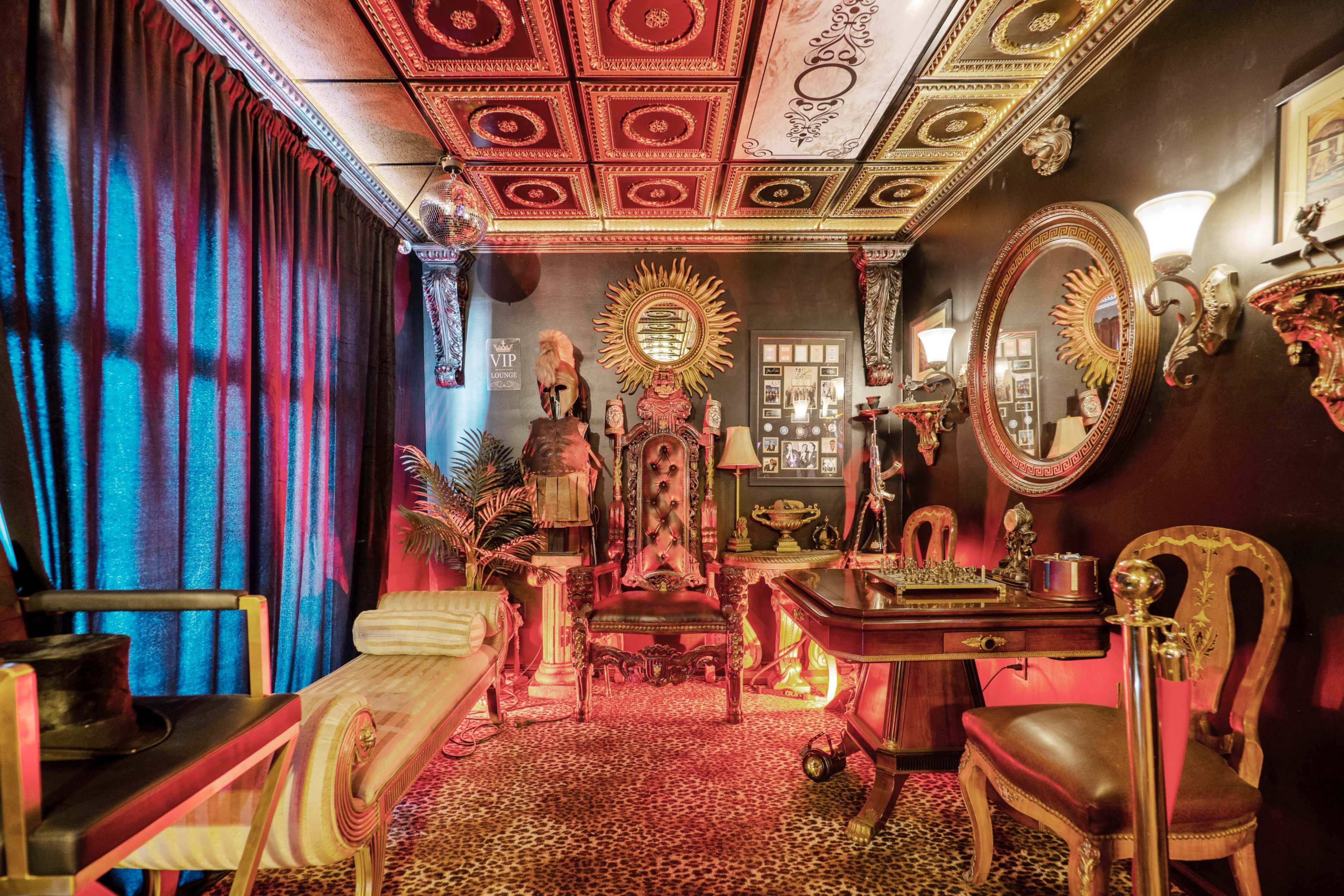 The room features an ornate chair, a wooden desk, and dramatic decor with rich textures and bold colors.