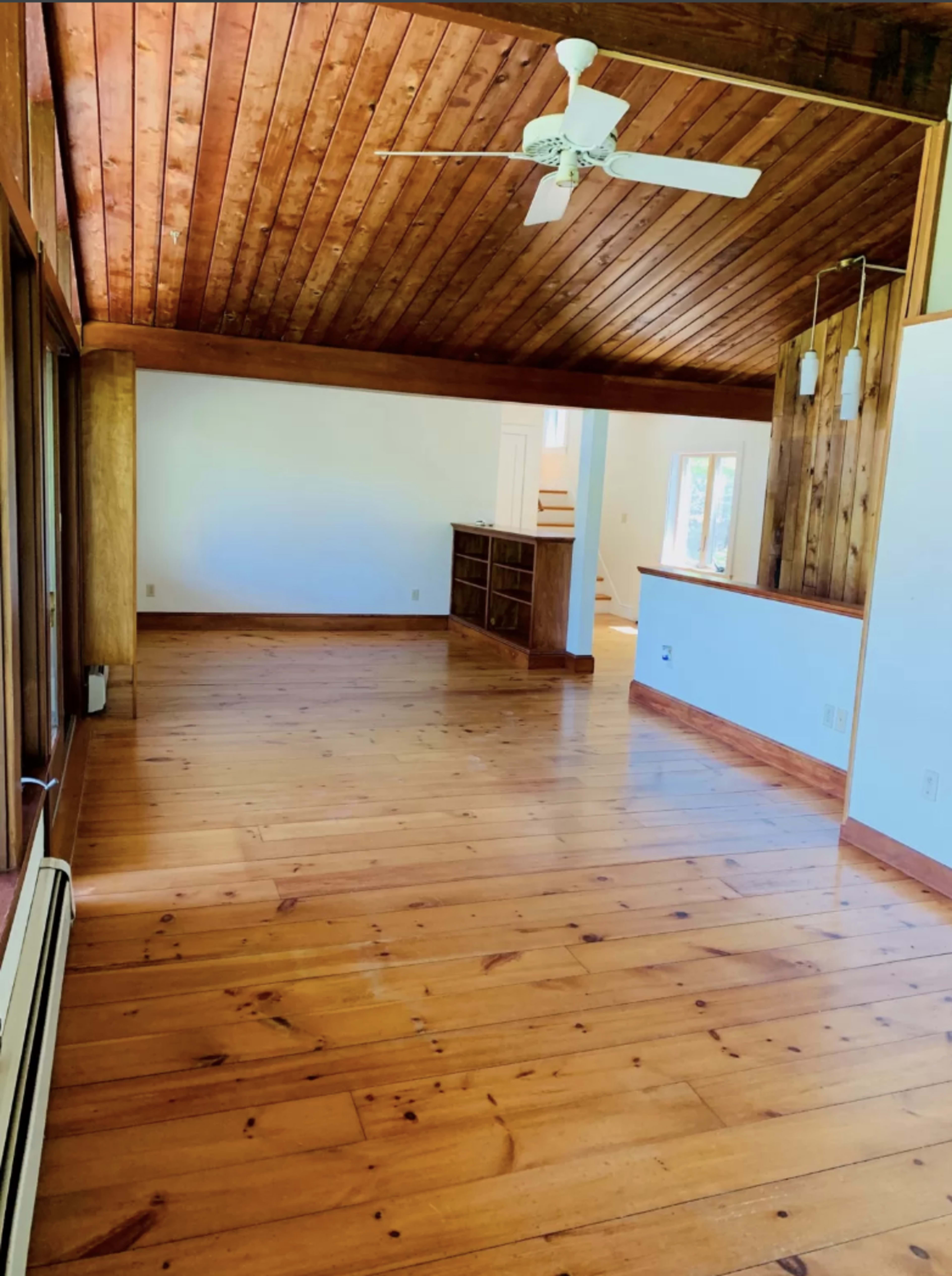 A spacious room with wooden flooring and a vaulted ceiling, featuring large windows and a ceiling fan.