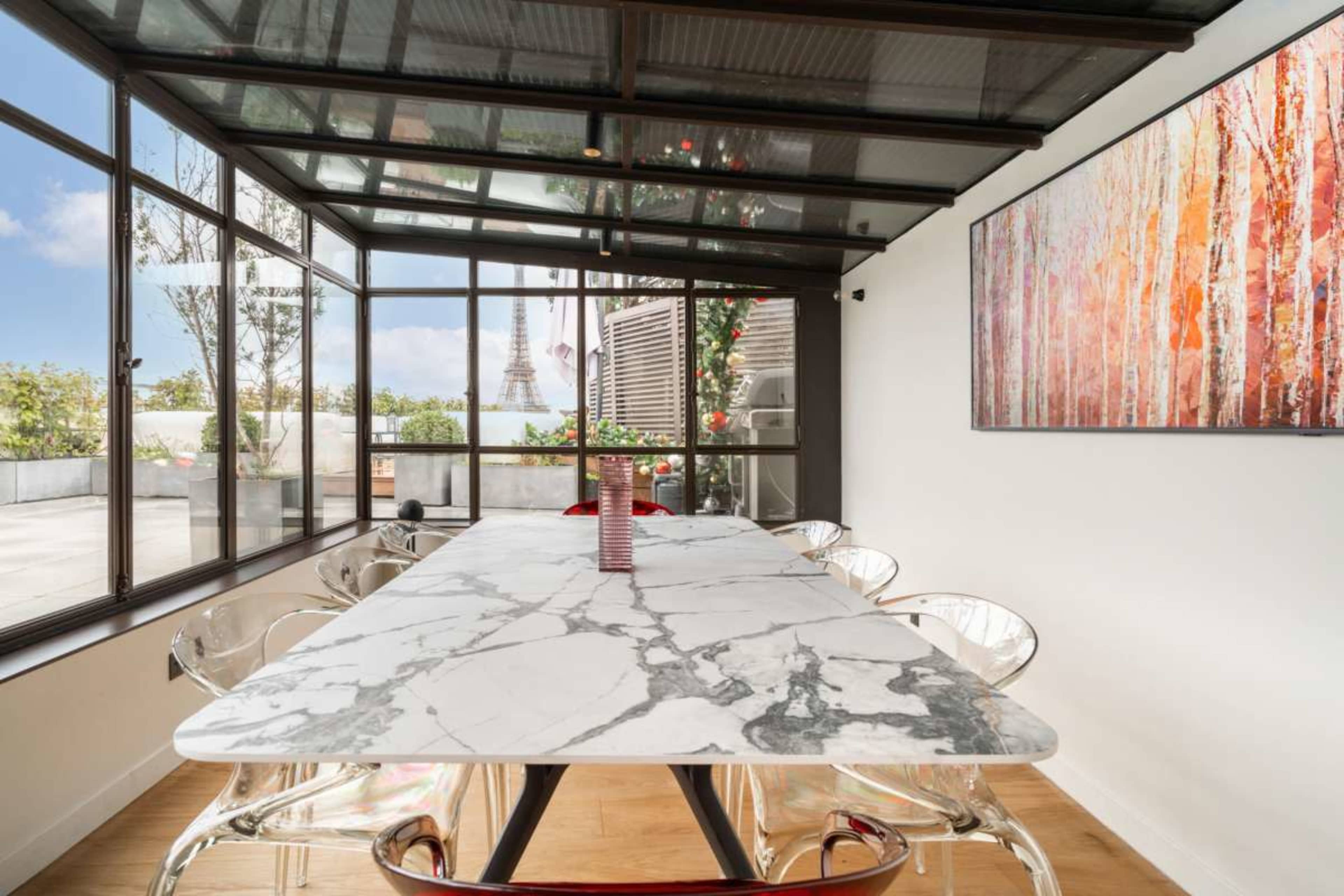 A modern dining area features a large marble table surrounded by transparent chairs, with large windows showcasing an outdoor view.