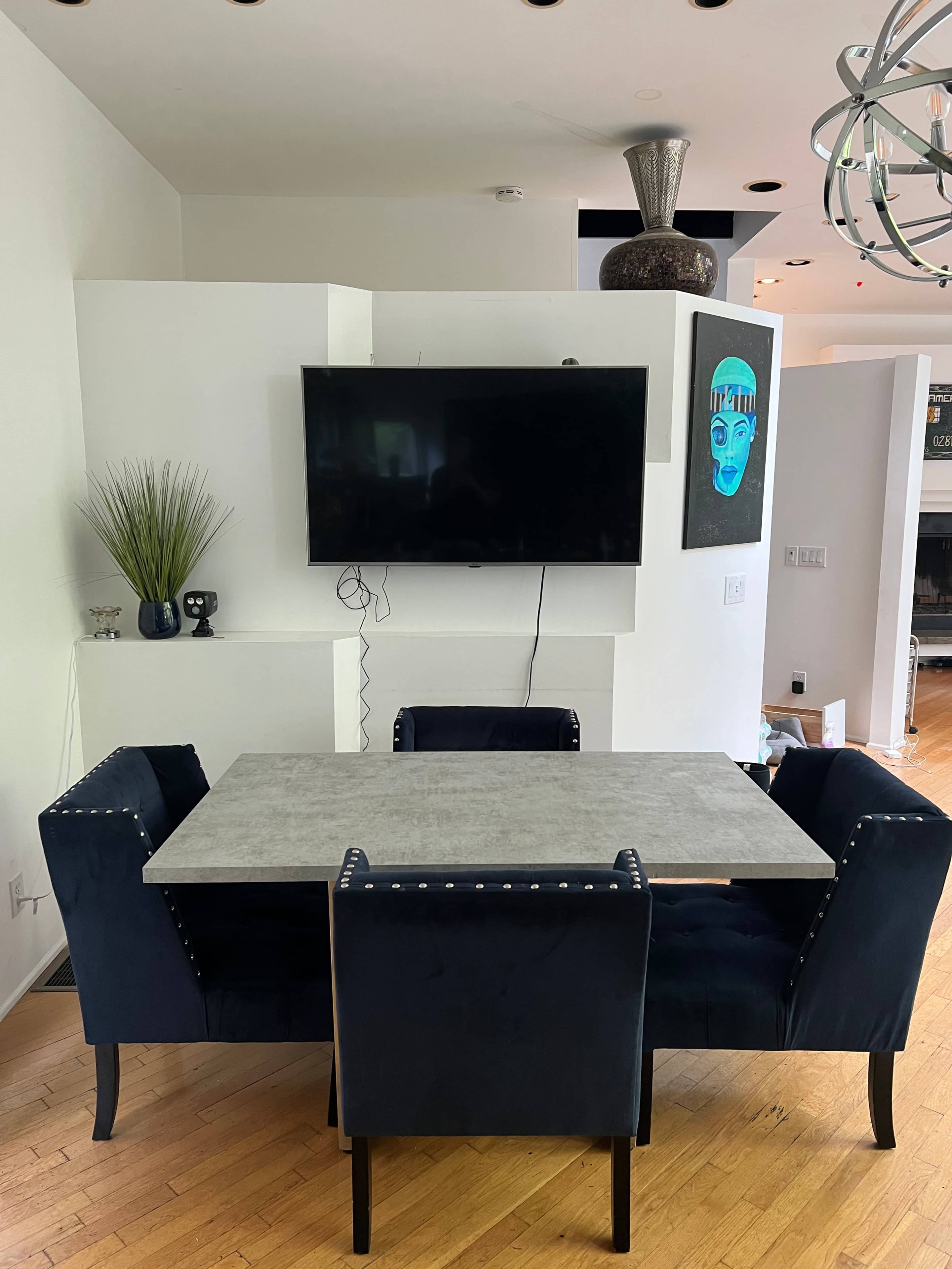 A modern dining area features a rectangular concrete table surrounded by four blue upholstered chairs, with a wall-mounted TV and artwork in the background.