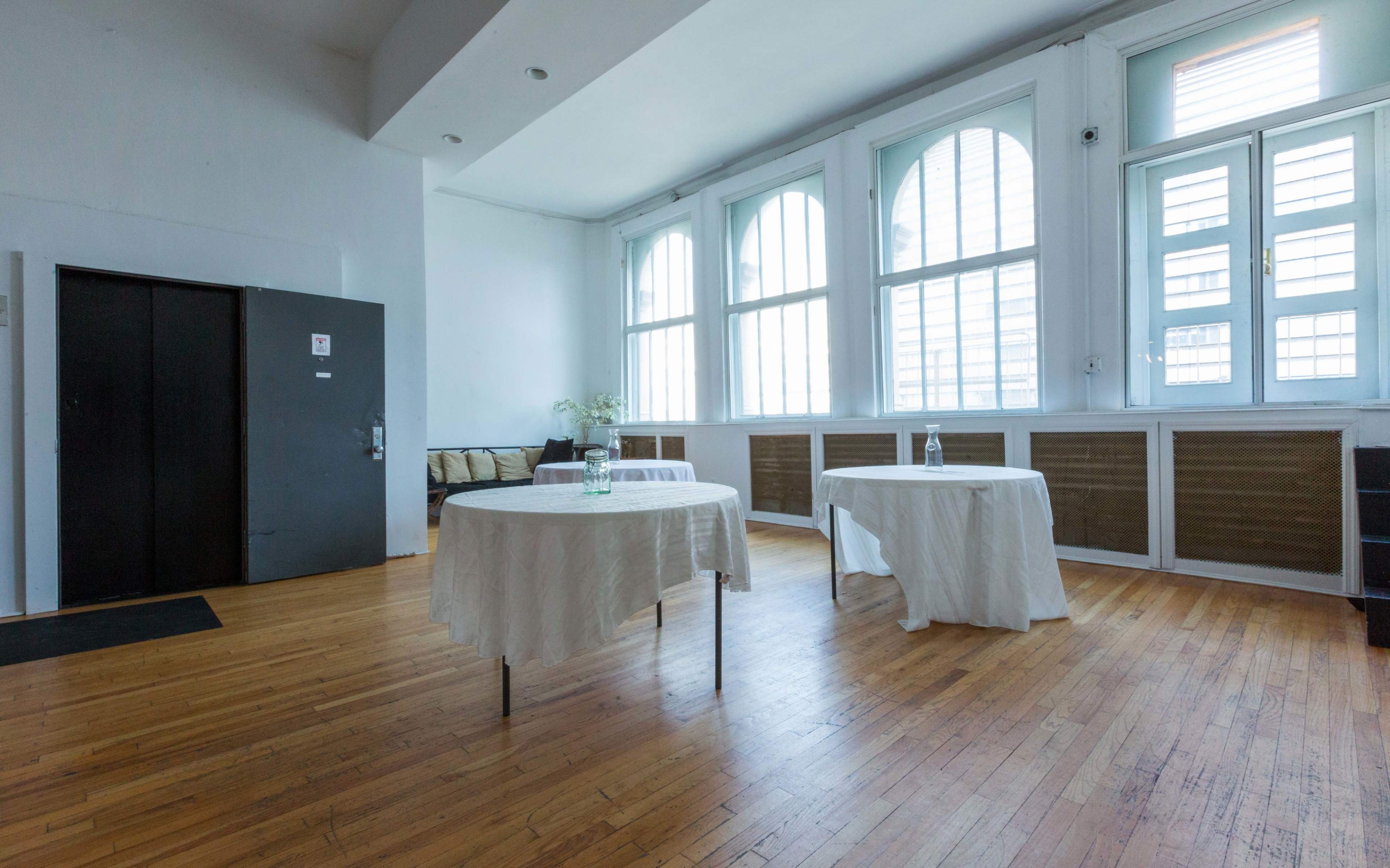The image shows a minimalist room with two small tables covered in white cloths, large windows letting in natural light, and an elevator door on the left.