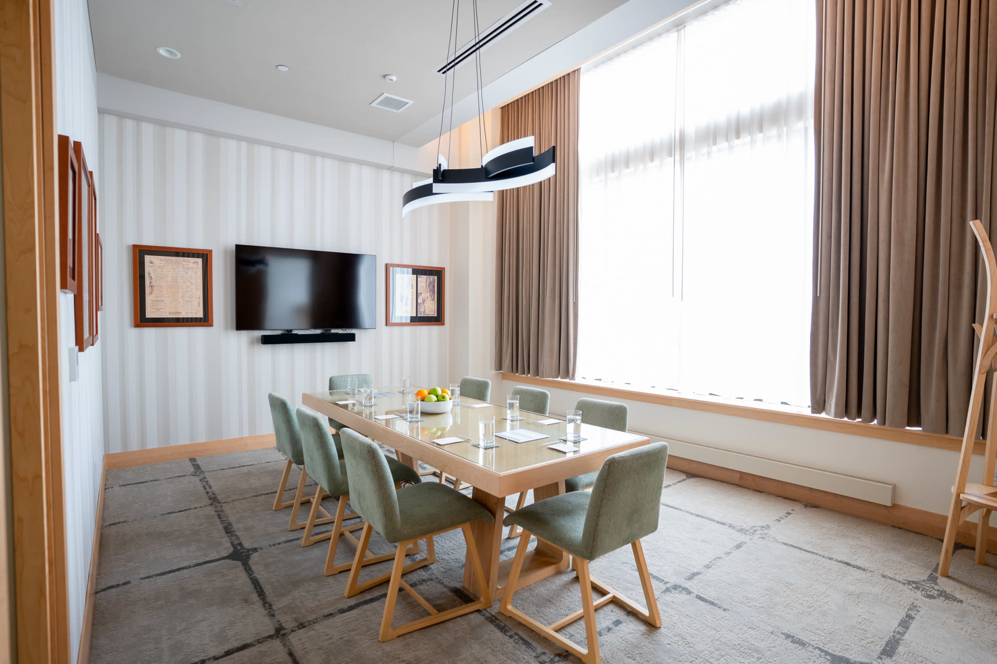 A conference room features a large wooden table surrounded by green chairs, with a television screen on the wall and large windows dressed with beige curtains.