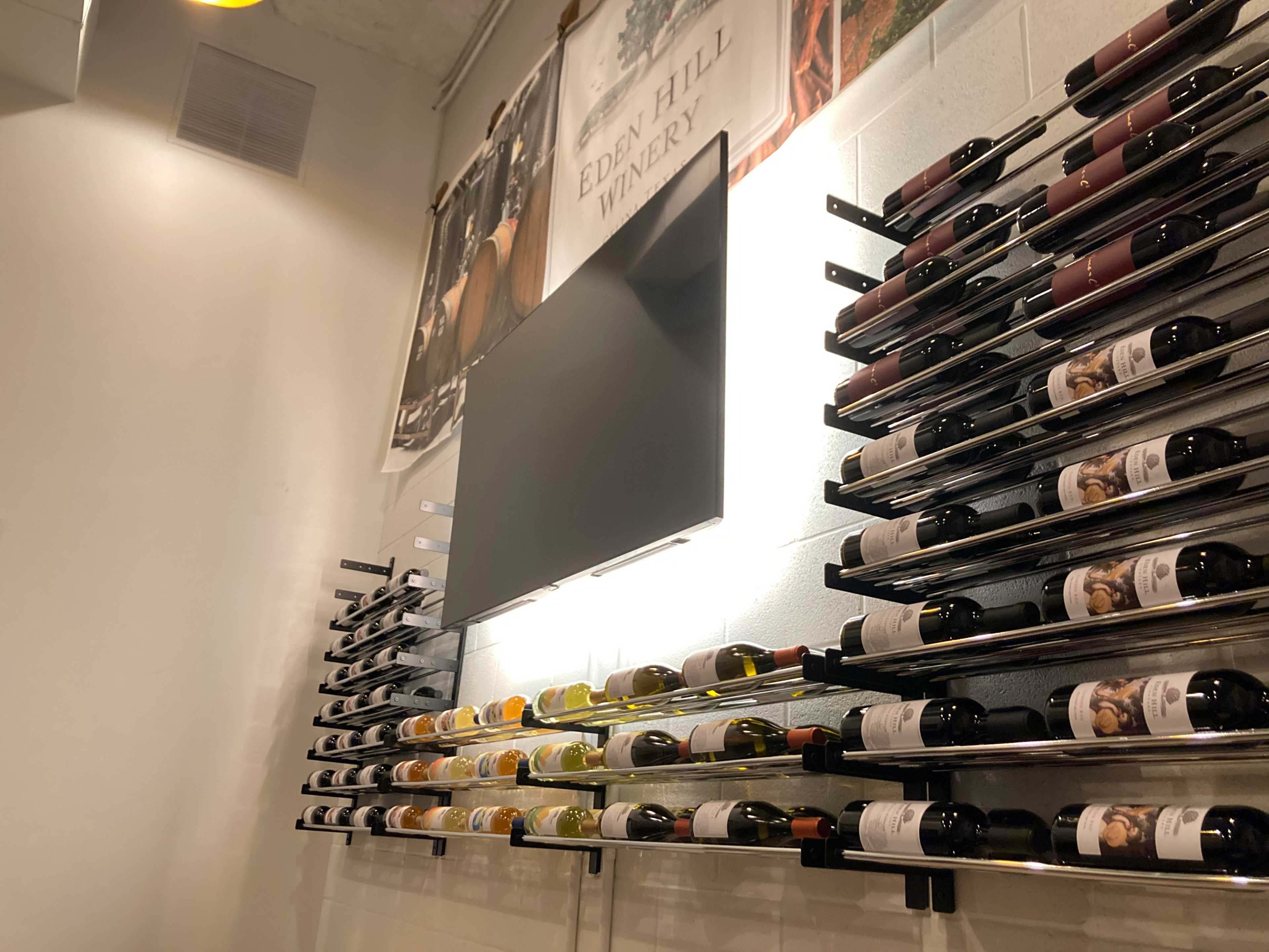 The image shows a wall-mounted wine rack displaying several bottles of wine alongside a flat-screen television and promotional material for Eden Hill Winery.