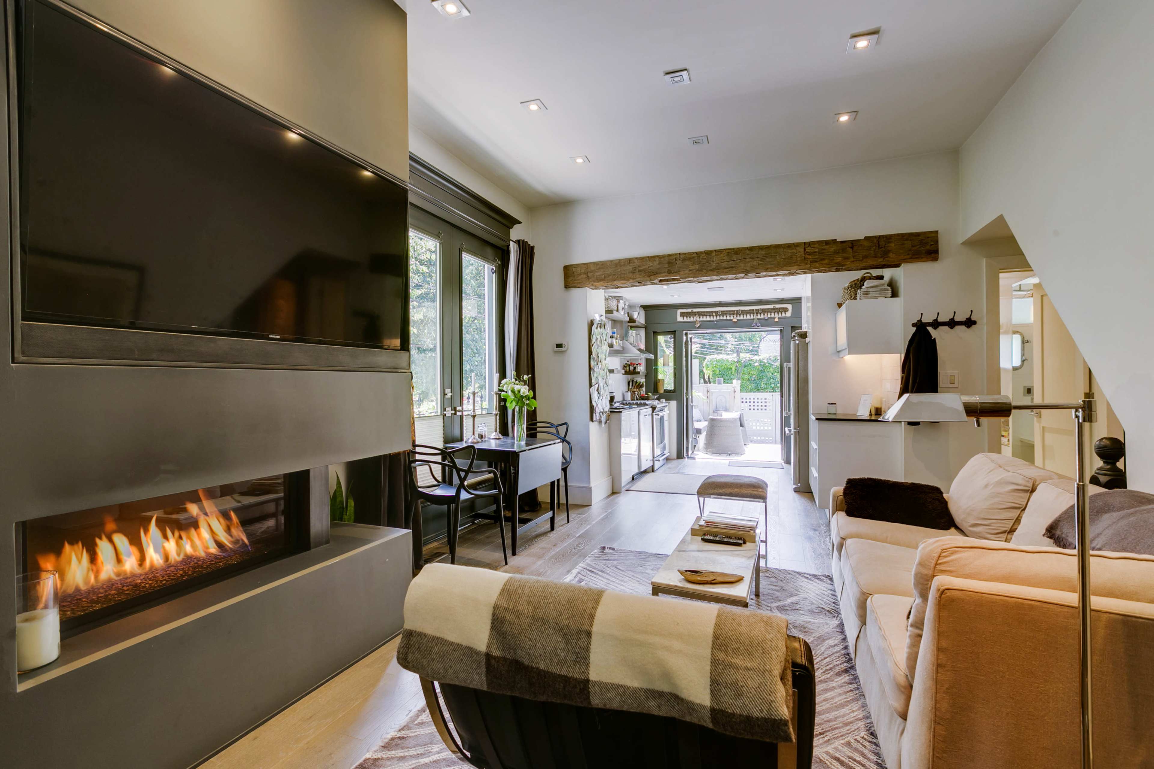 A modern living room with a flat-screen TV mounted on a wall, a fireplace below, and an open view into a well-lit kitchen area.
