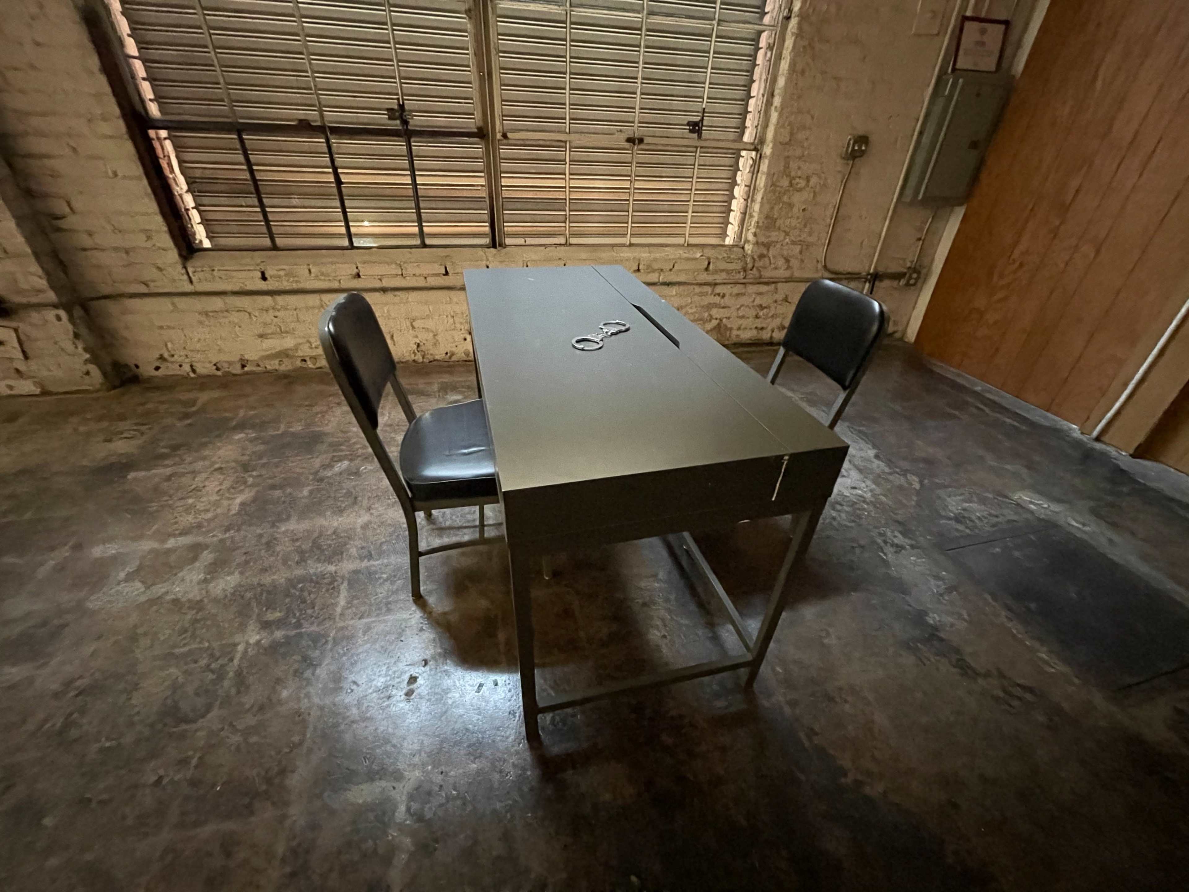 A metal table with two chairs is positioned on a concrete floor under a window with blinds partially pulled down.