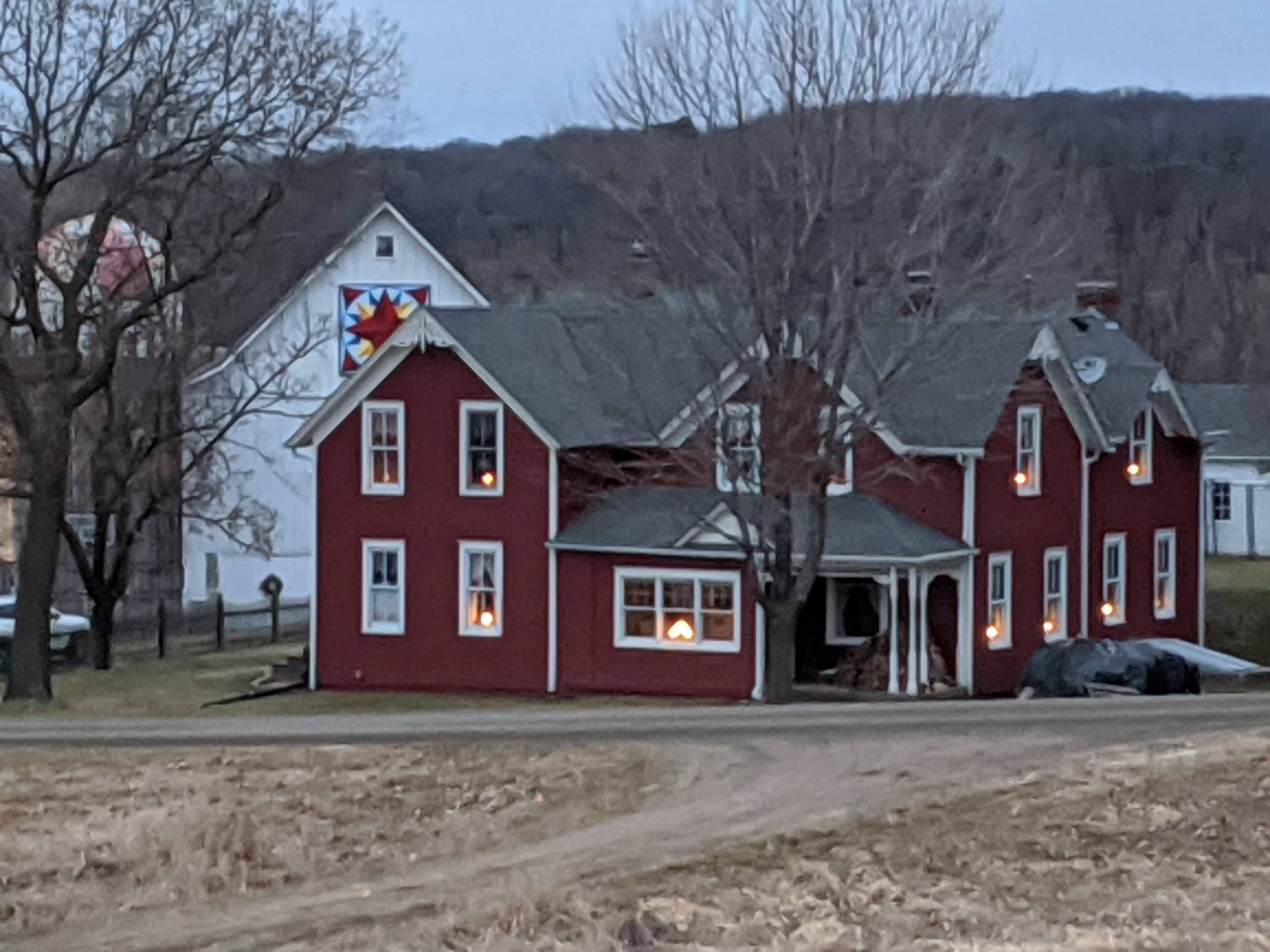 A red farmhouse with illuminated windows and a quilt pattern on the adjoining white building is set against a backdrop of bare trees and a twilight sky.