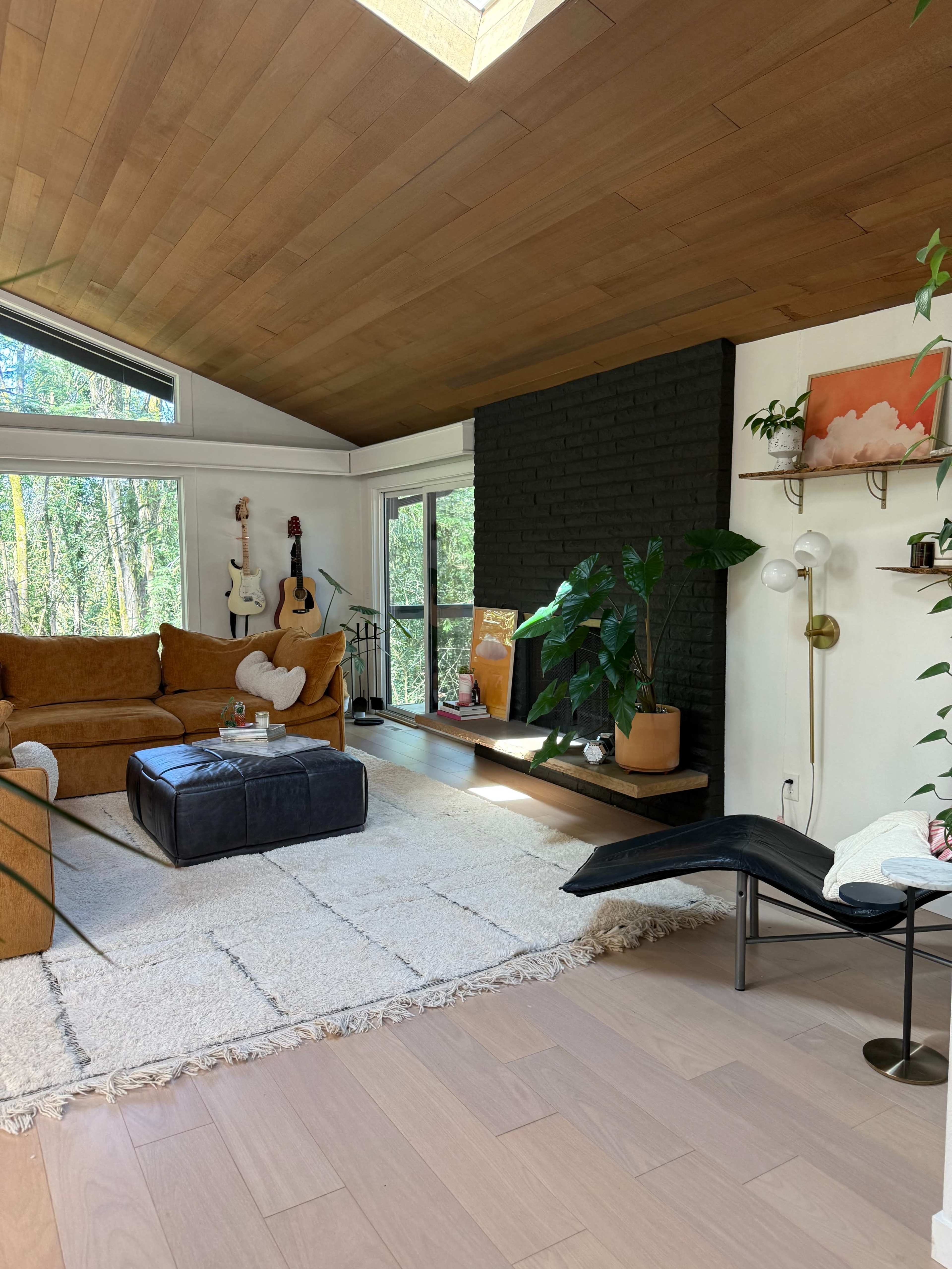 A cozy living room features a brown sofa, a black ottoman, plants, and large windows overlooking a forested area.