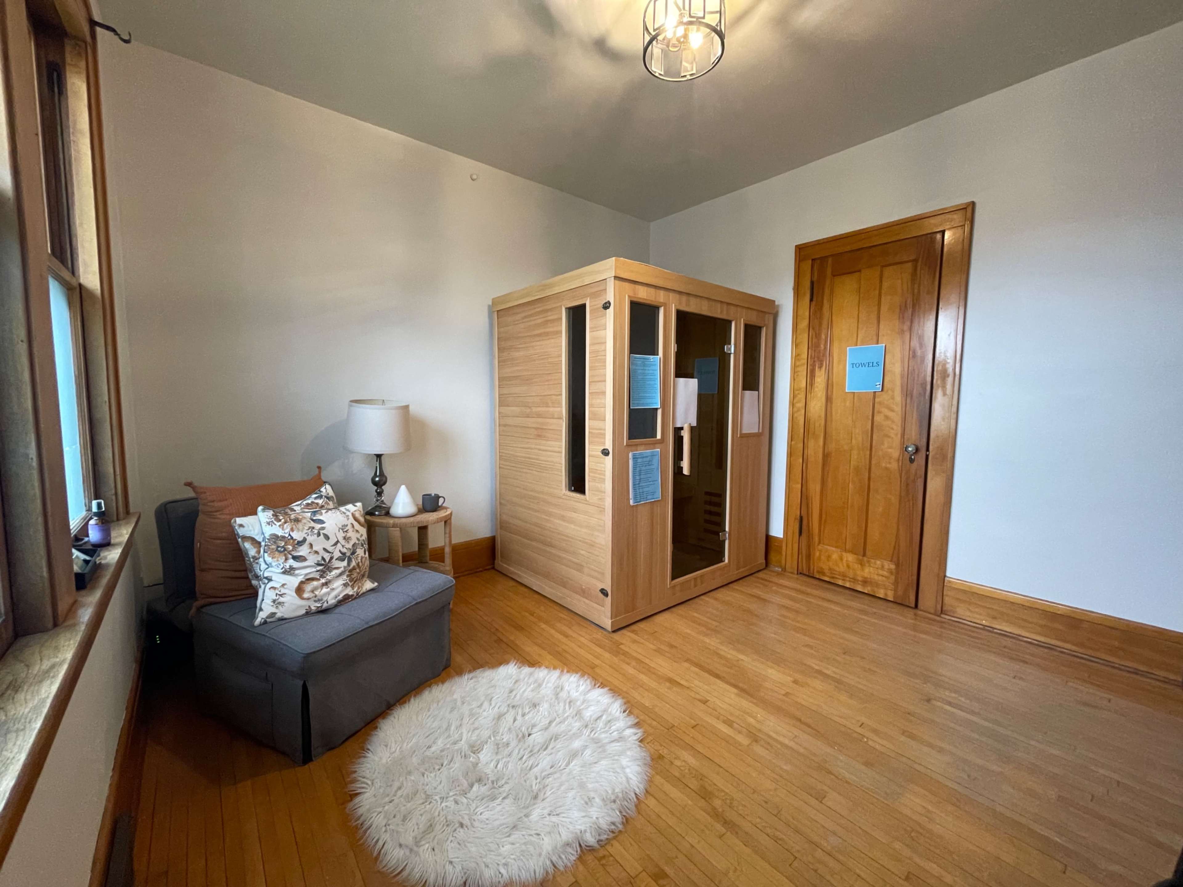 A spacious room with wooden floors, featuring a gray armchair beside a small lamp and a light-colored rug, along with a wooden door and a sauna in the corner.