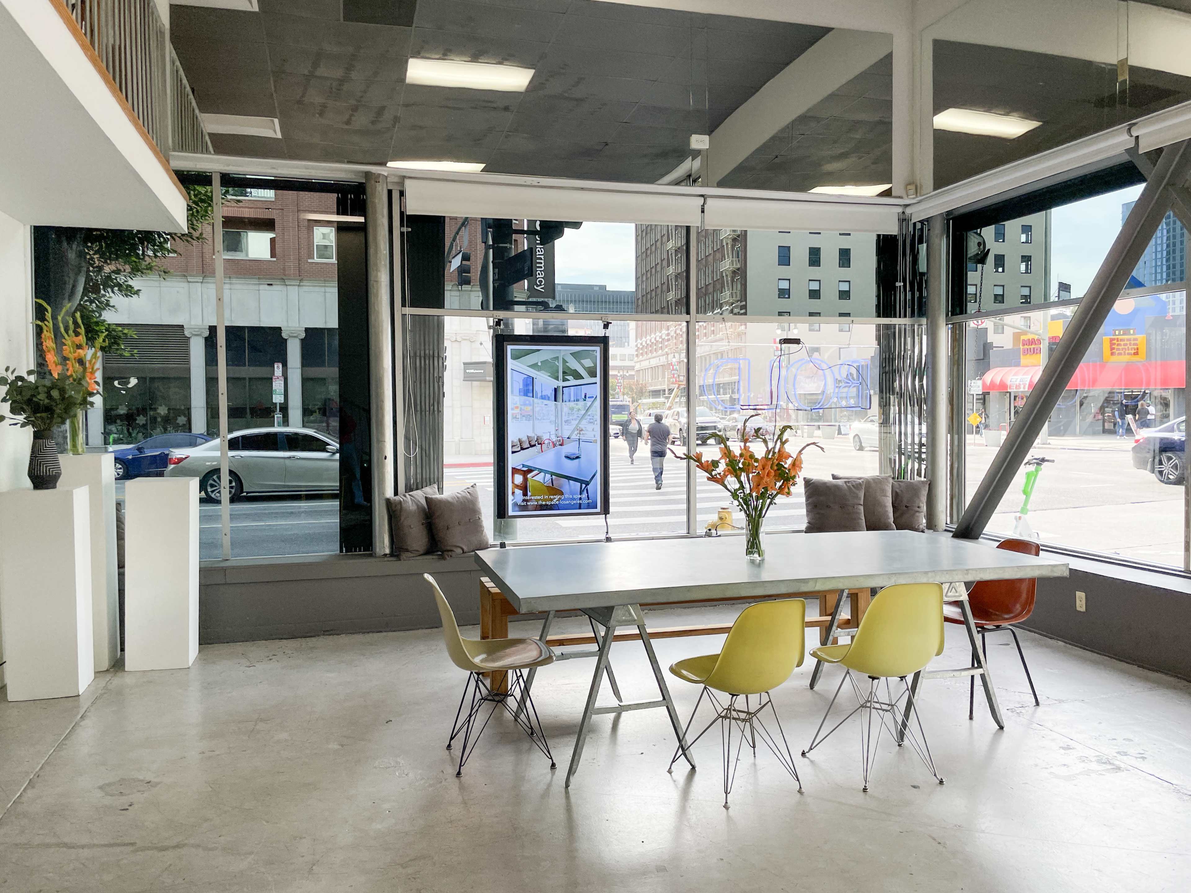 A spacious indoor setting features a large table surrounded by several chairs, with large windows offering a view of a city street.