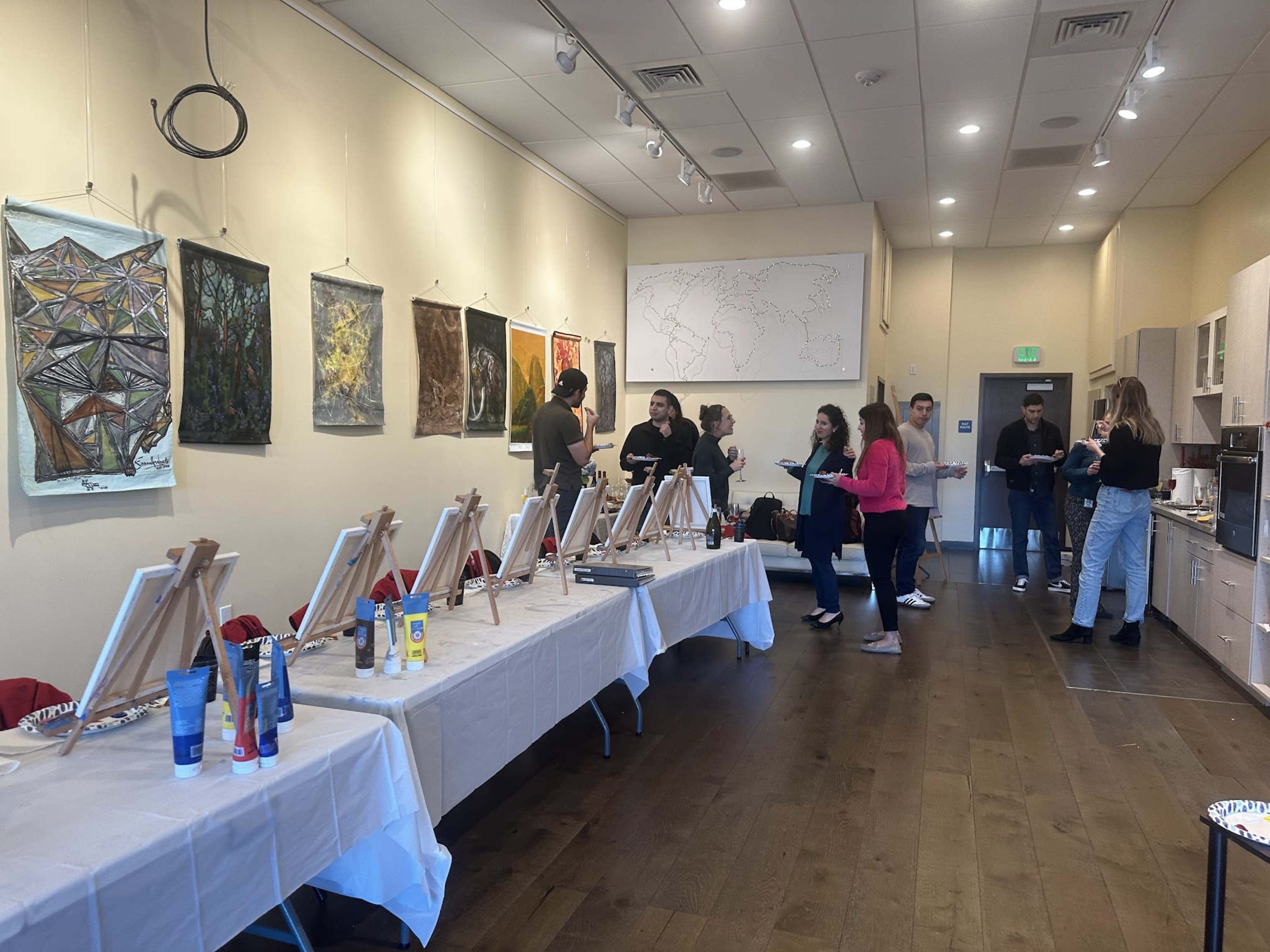 A group of people is gathered in a studio environment with easels set up on tables, canvases ready for painting, and various art supplies displayed.