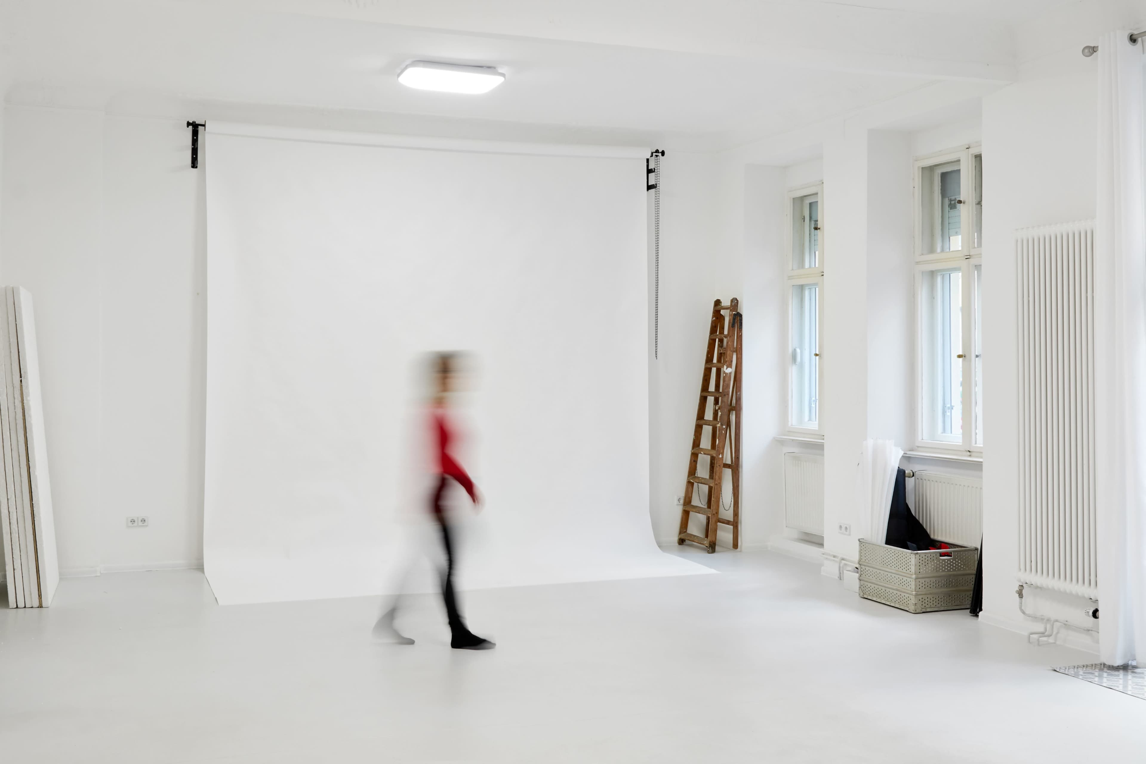 A person walks past a blank white backdrop in a bright studio with large windows and a wooden ladder.