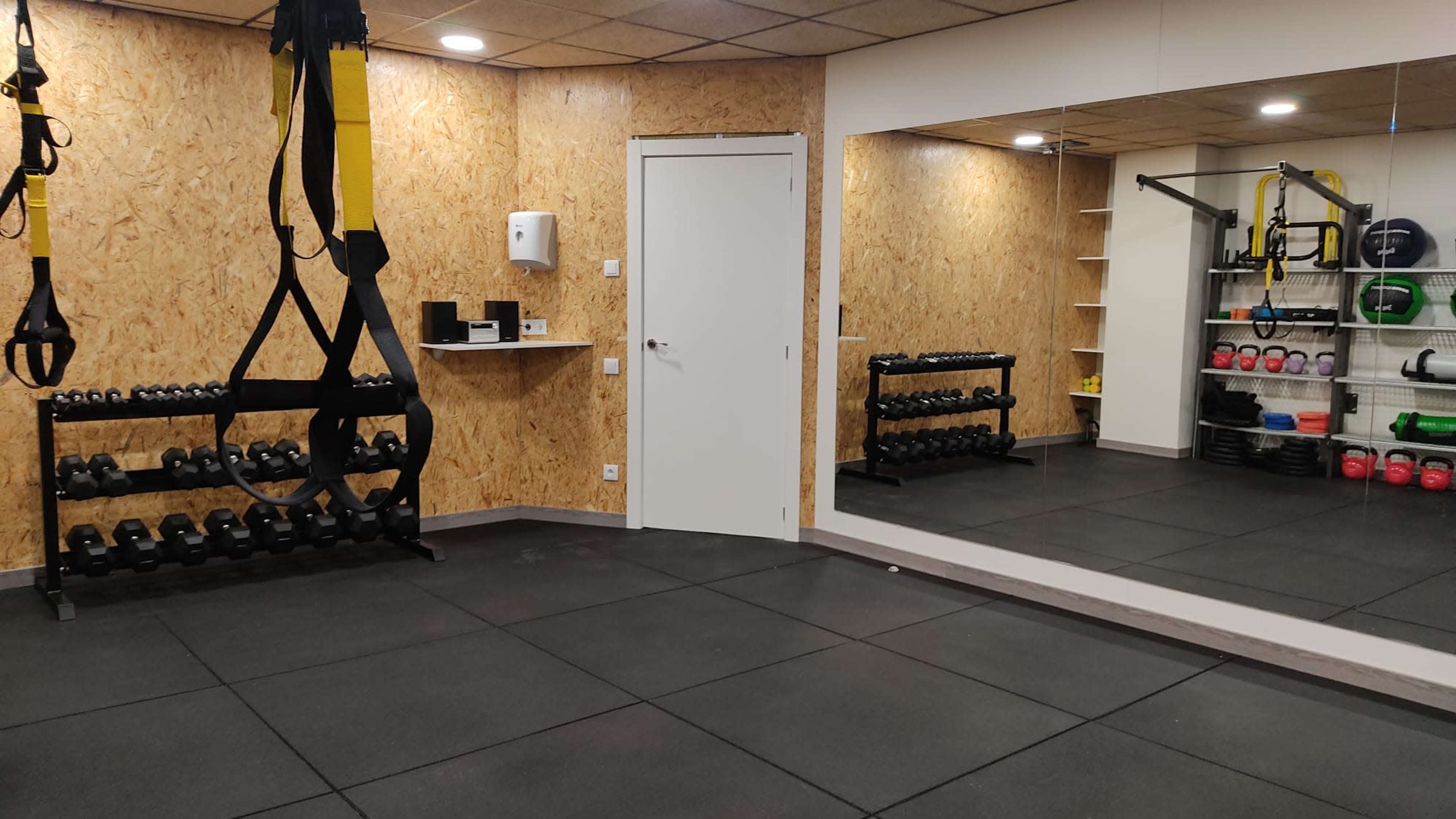 The image shows a fitness room with a mirrored wall, a rack of dumbbells, and various workout equipment arranged on shelves.
