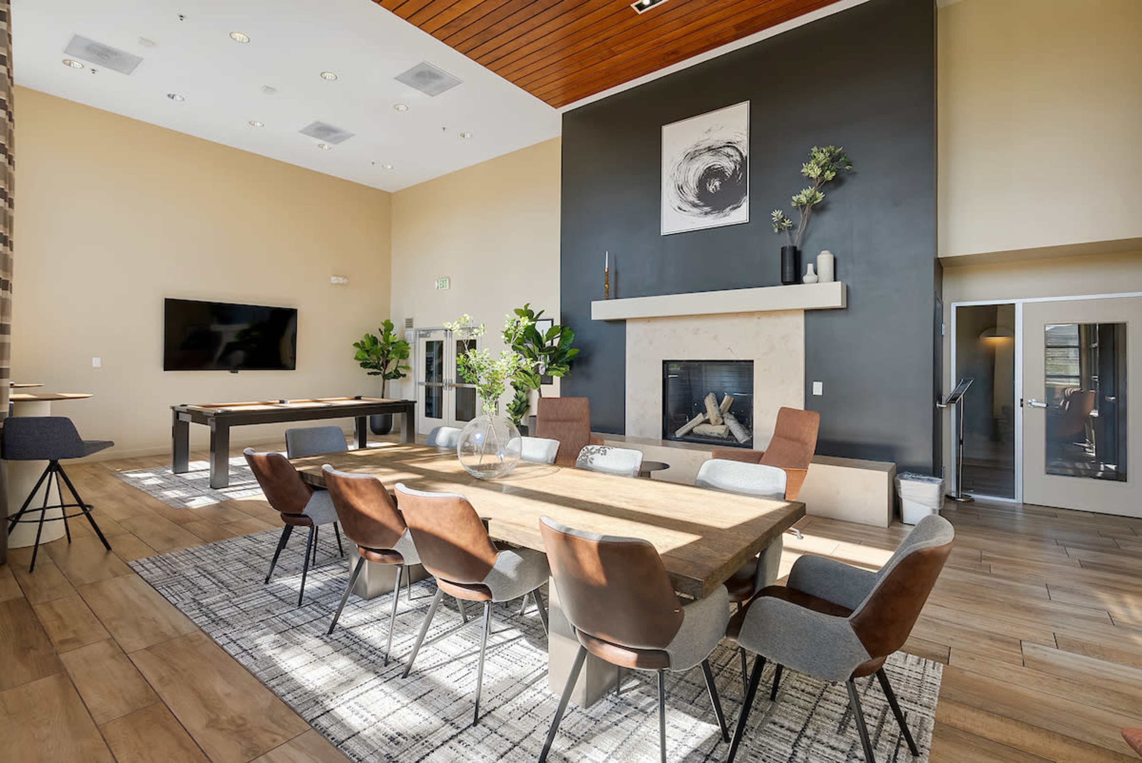 The image shows a modern communal room with a large wooden dining table surrounded by various chairs, a fireplace, a television, and potted plants.