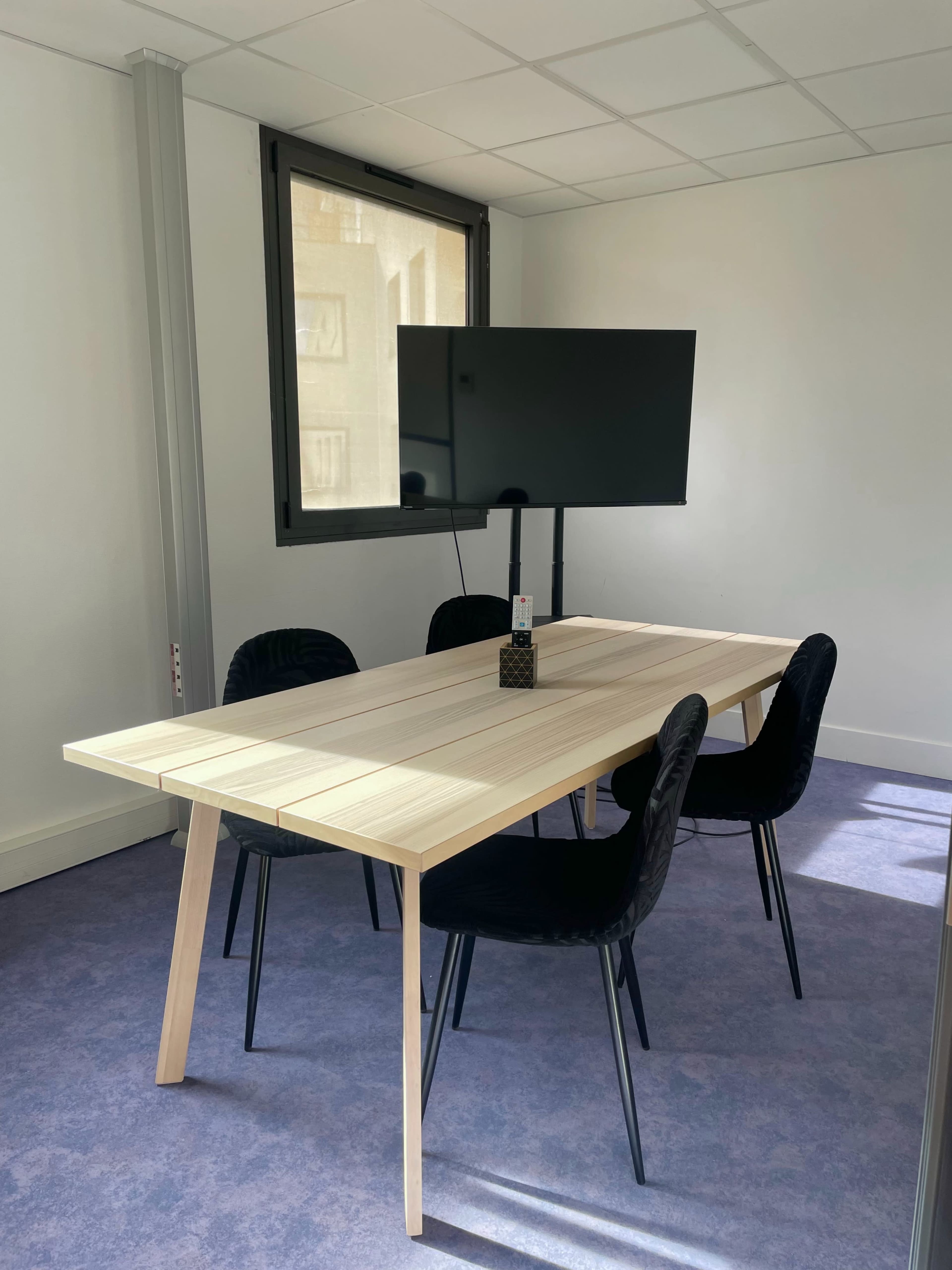 A simple meeting room with a rectangular wooden table and four black chairs, accompanied by a wall-mounted television.
