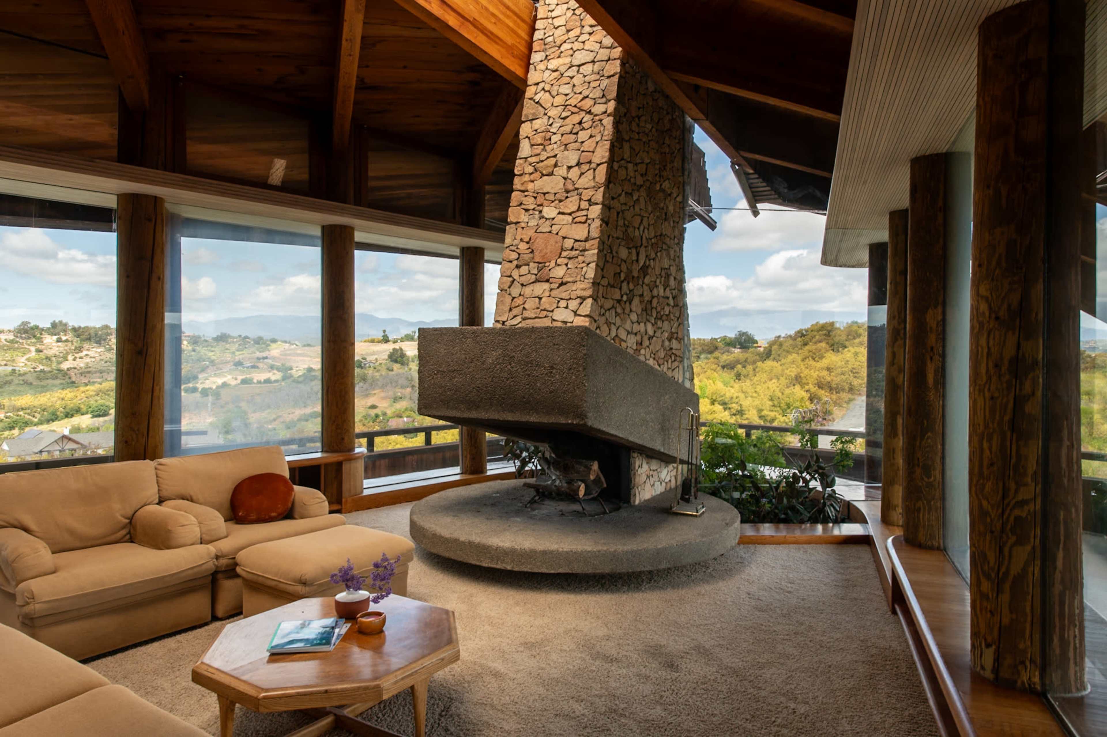 The image shows a spacious living room with a large stone fireplace at its center, surrounded by windows that offer views of the landscape outside.