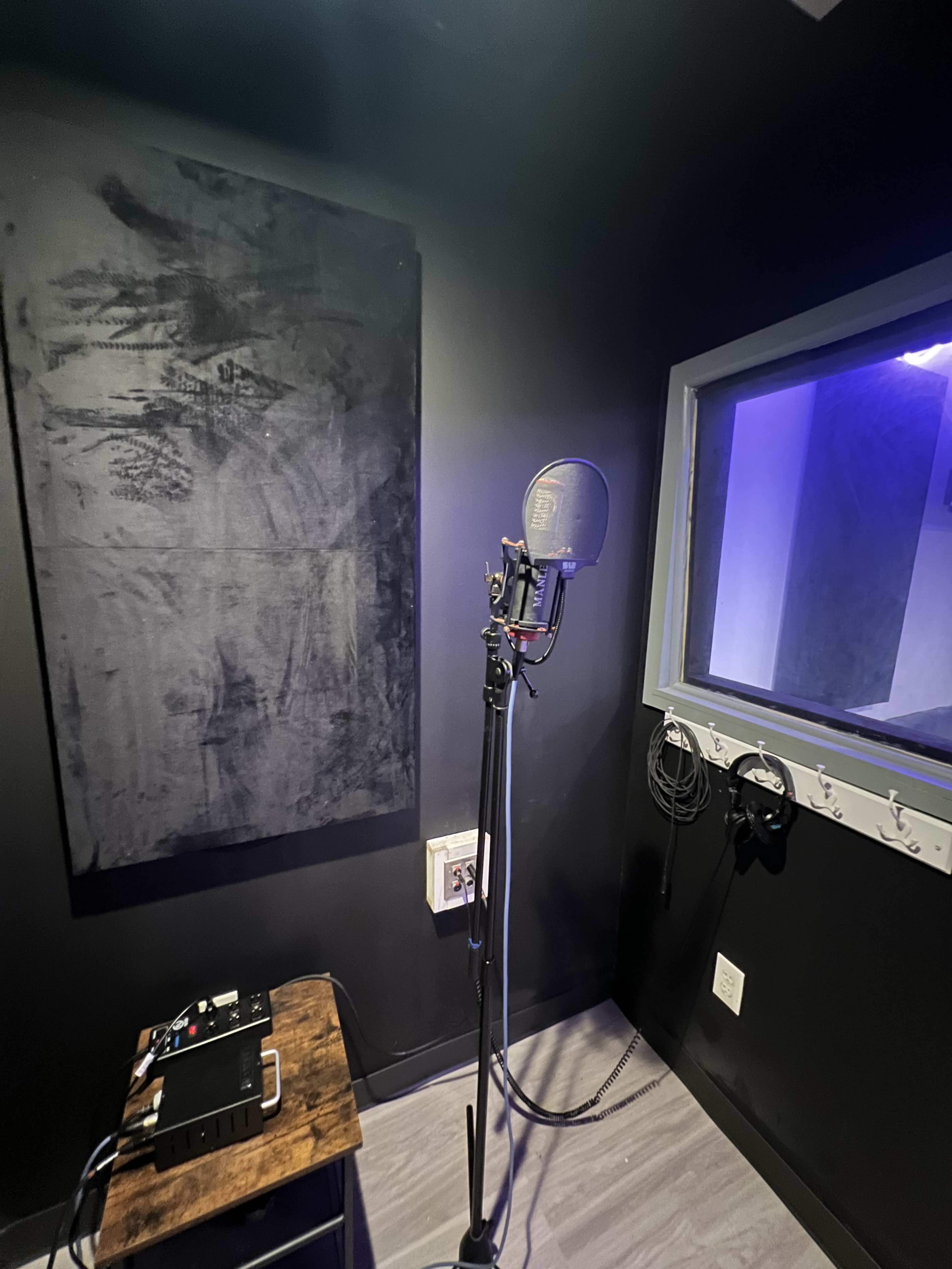 The image shows a soundproof recording booth with a microphone on a stand and audio equipment on a wooden table.