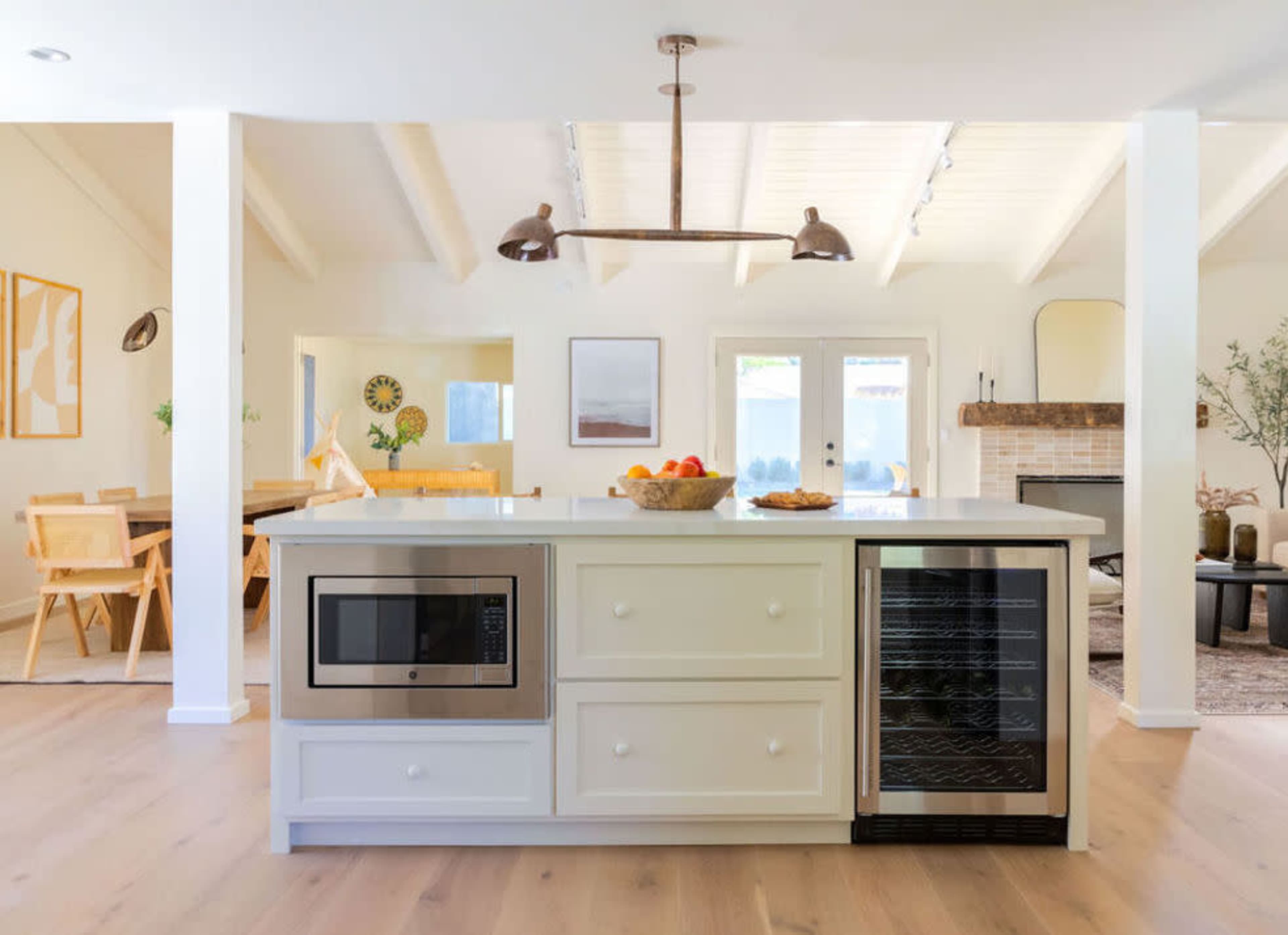 A modern kitchen features a central island with a microwave and wine fridge, surrounded by bright, airy spaces and wooden furnishings.