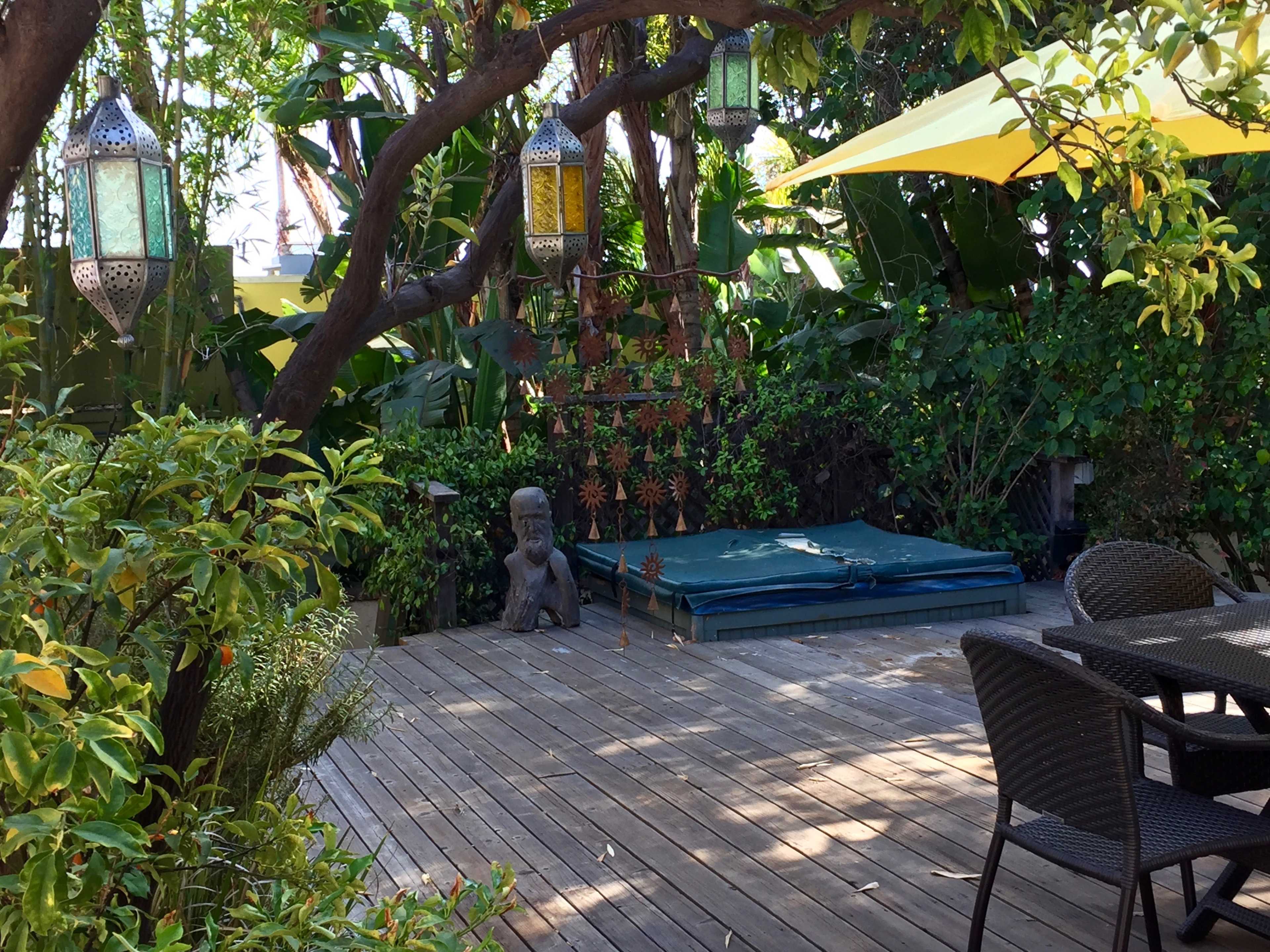 The image shows a wooden deck surrounded by greenery, featuring hanging lanterns and a statue, with yellow awnings providing shade over patio furniture.