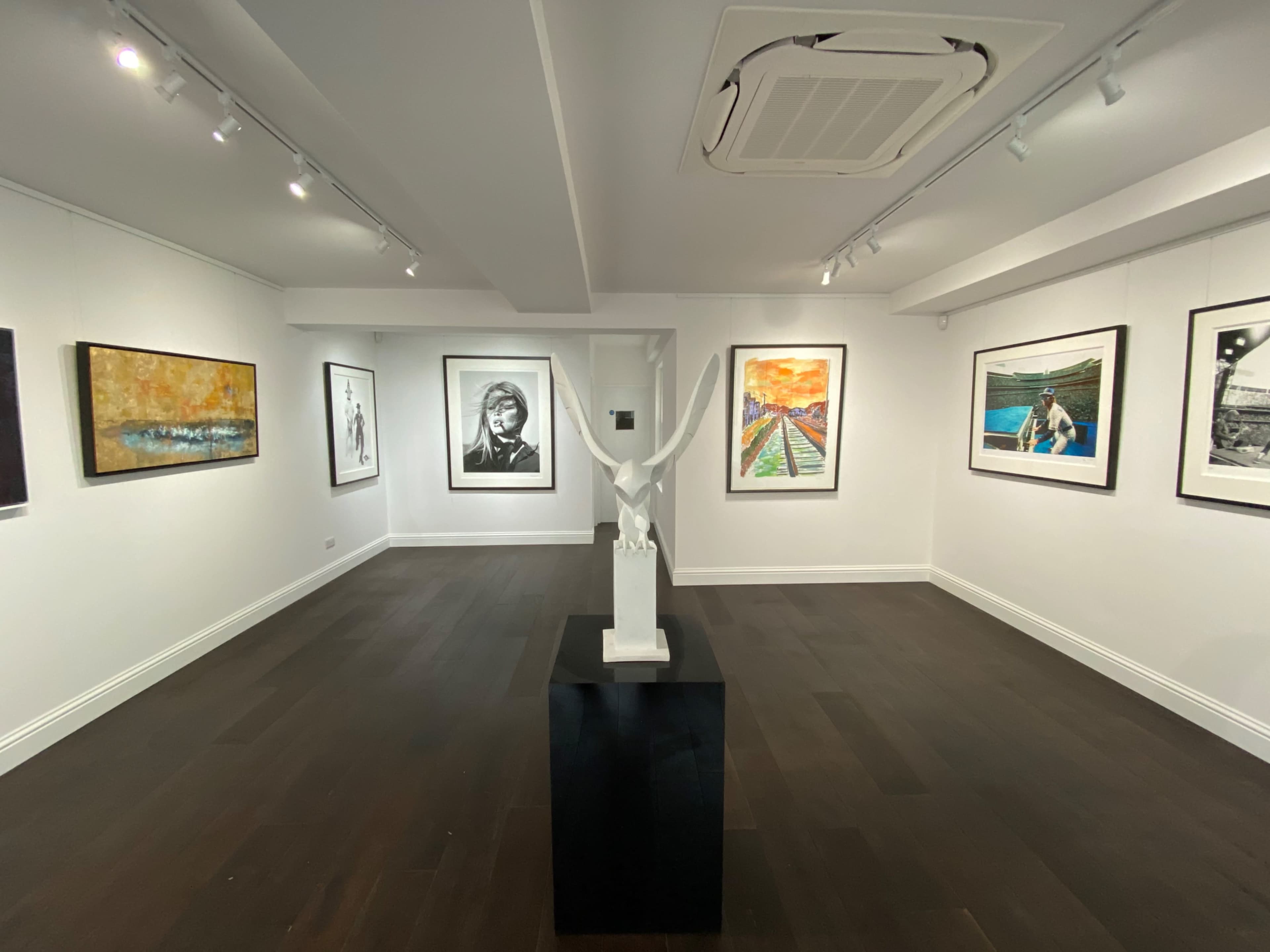 The image shows an art gallery interior with various framed artworks displayed on the walls and a white sculpture on a pedestal in the center.