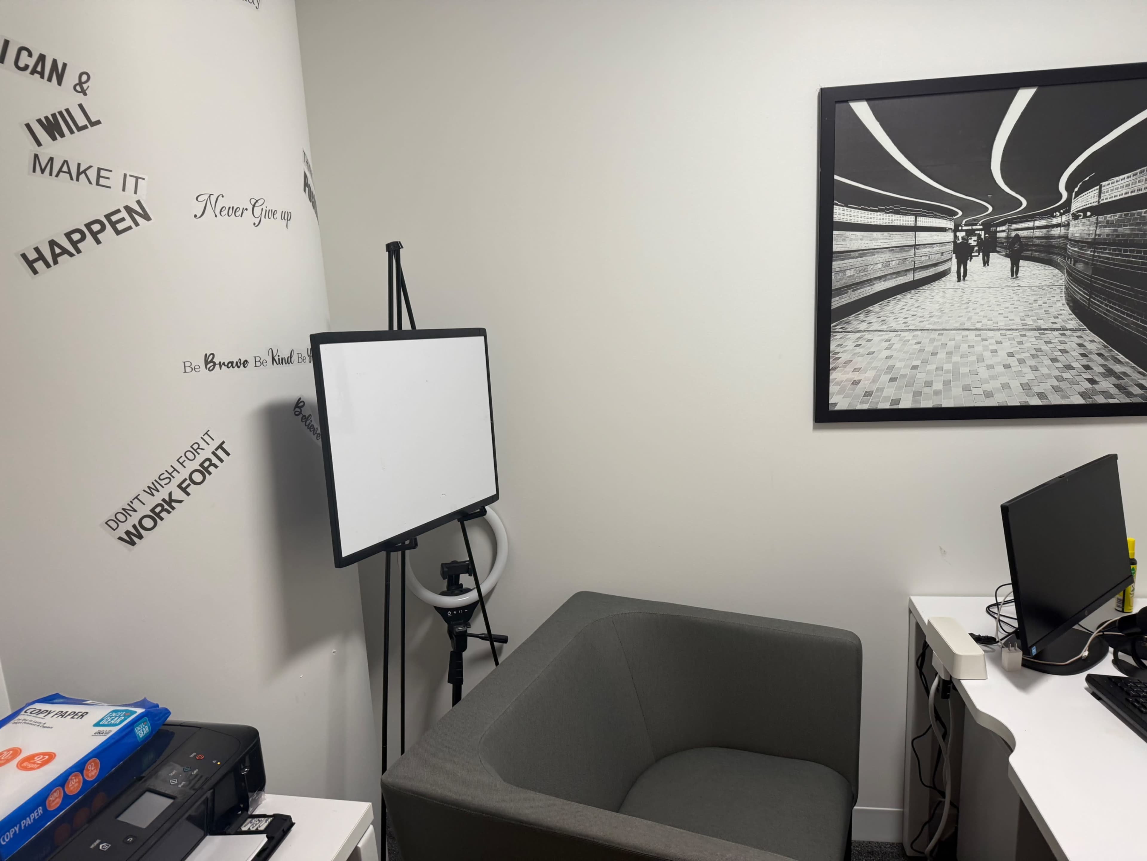The image shows a small office space featuring a gray chair, a whiteboard on an easel, a printer, and a desk with a computer.