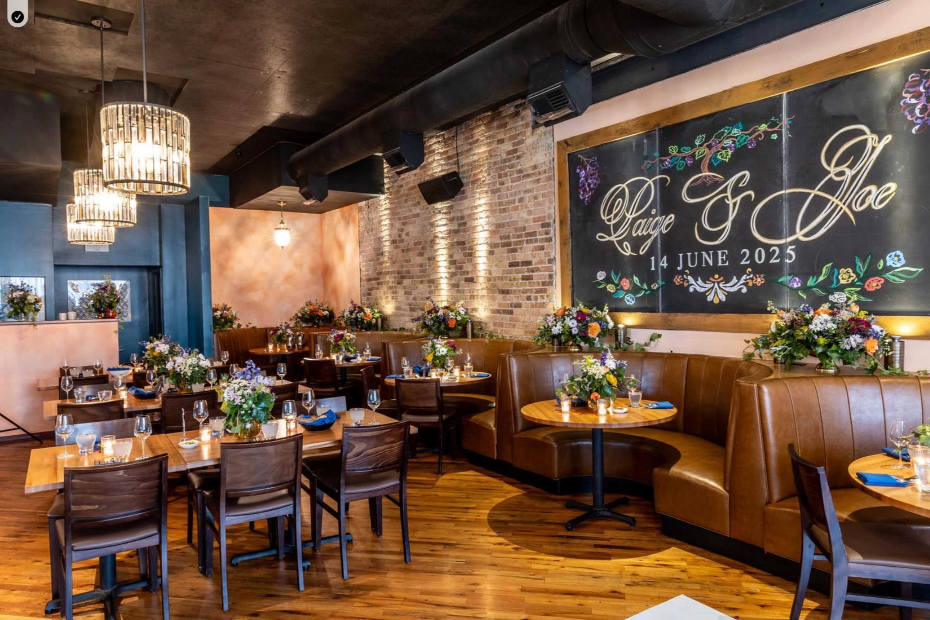 The image shows a modern restaurant interior with wooden booths, tables set for dining, and floral arrangements adorning the space.
