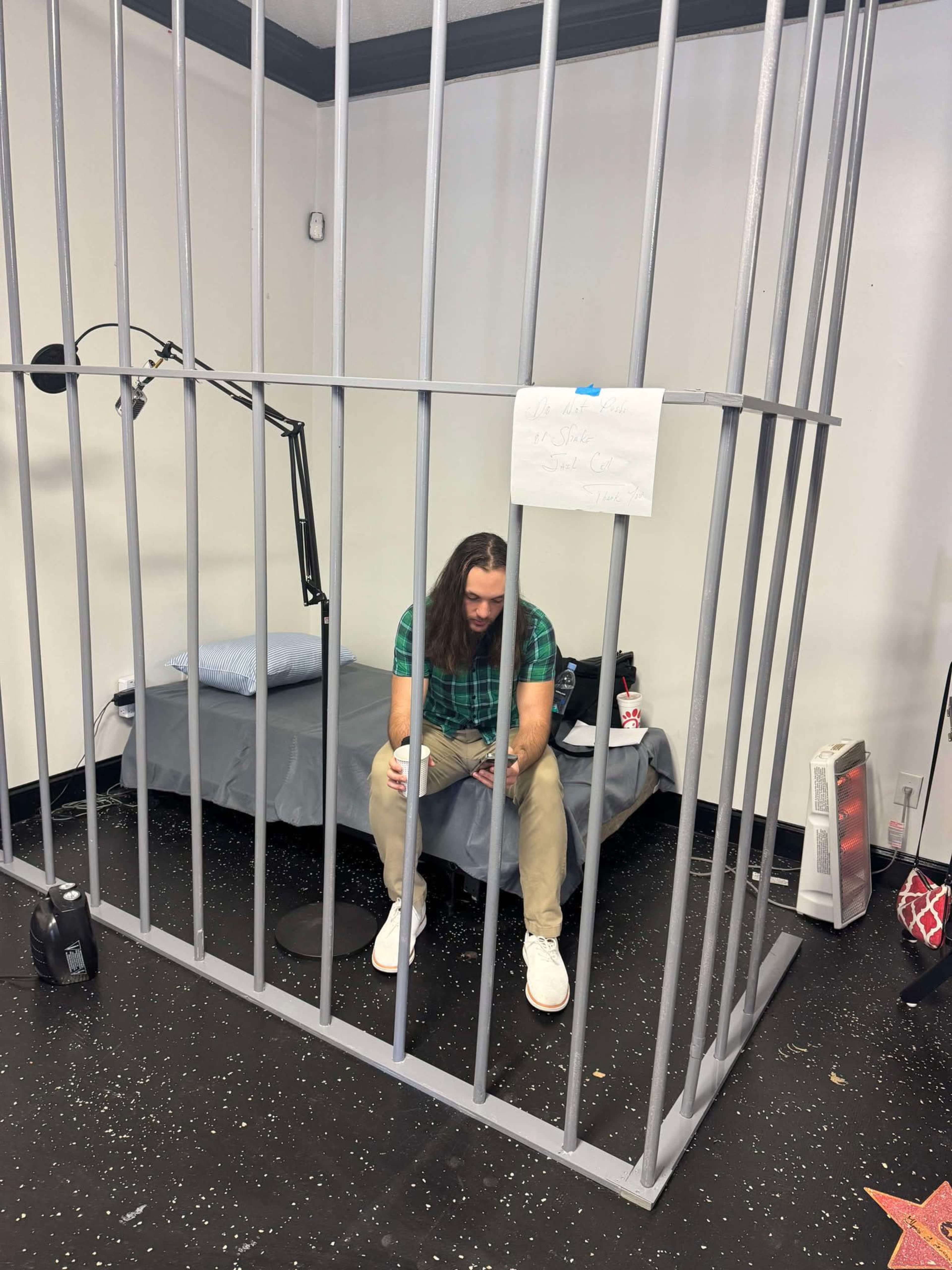 A person with long hair sits inside a cage-like structure, looking down at an object in their hand, with a bed and a lamp inside the enclosure.