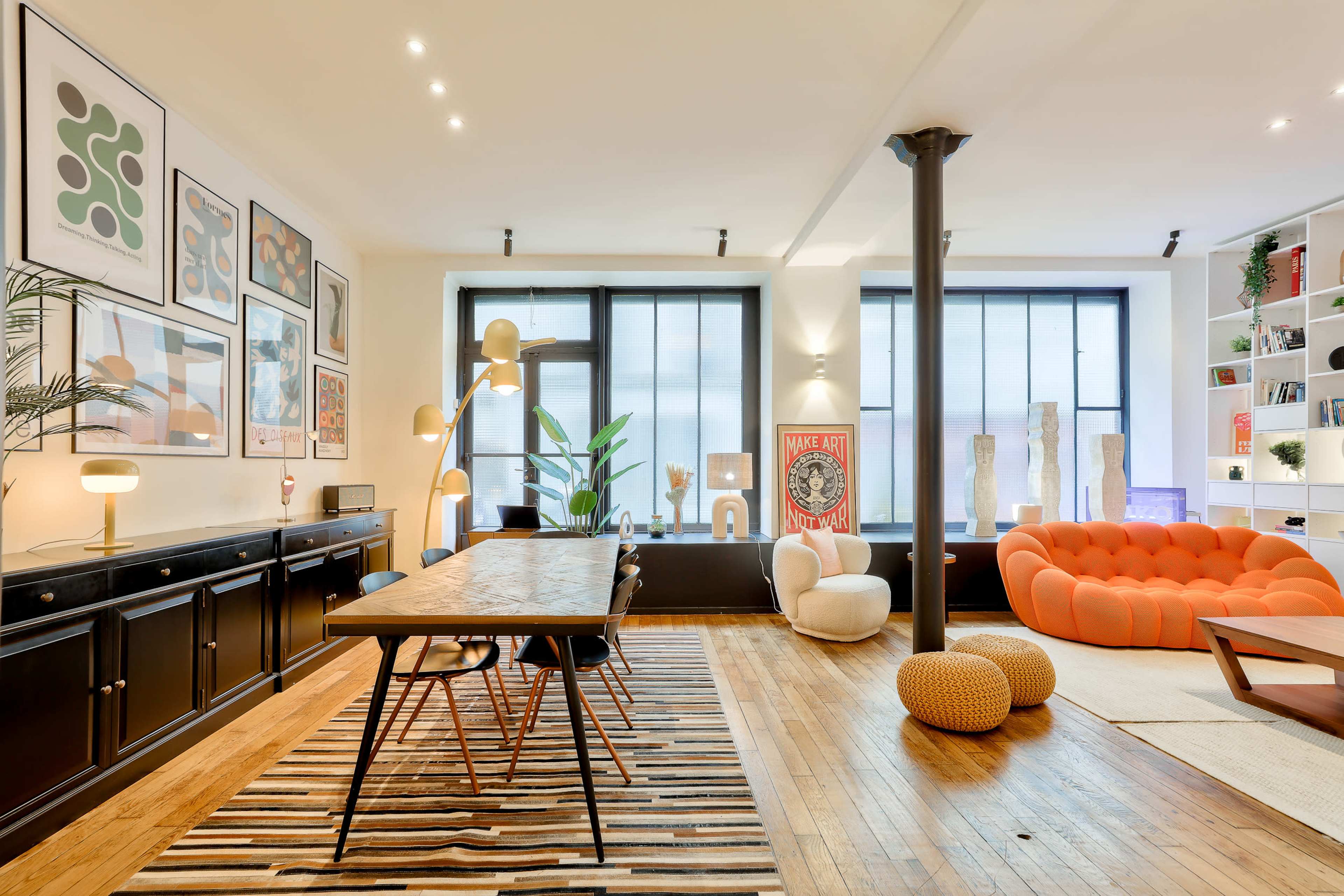 A modern living space featuring a dining area with a wooden table and chairs, a vibrant orange sofa, large windows, and various pieces of art displayed on the walls.