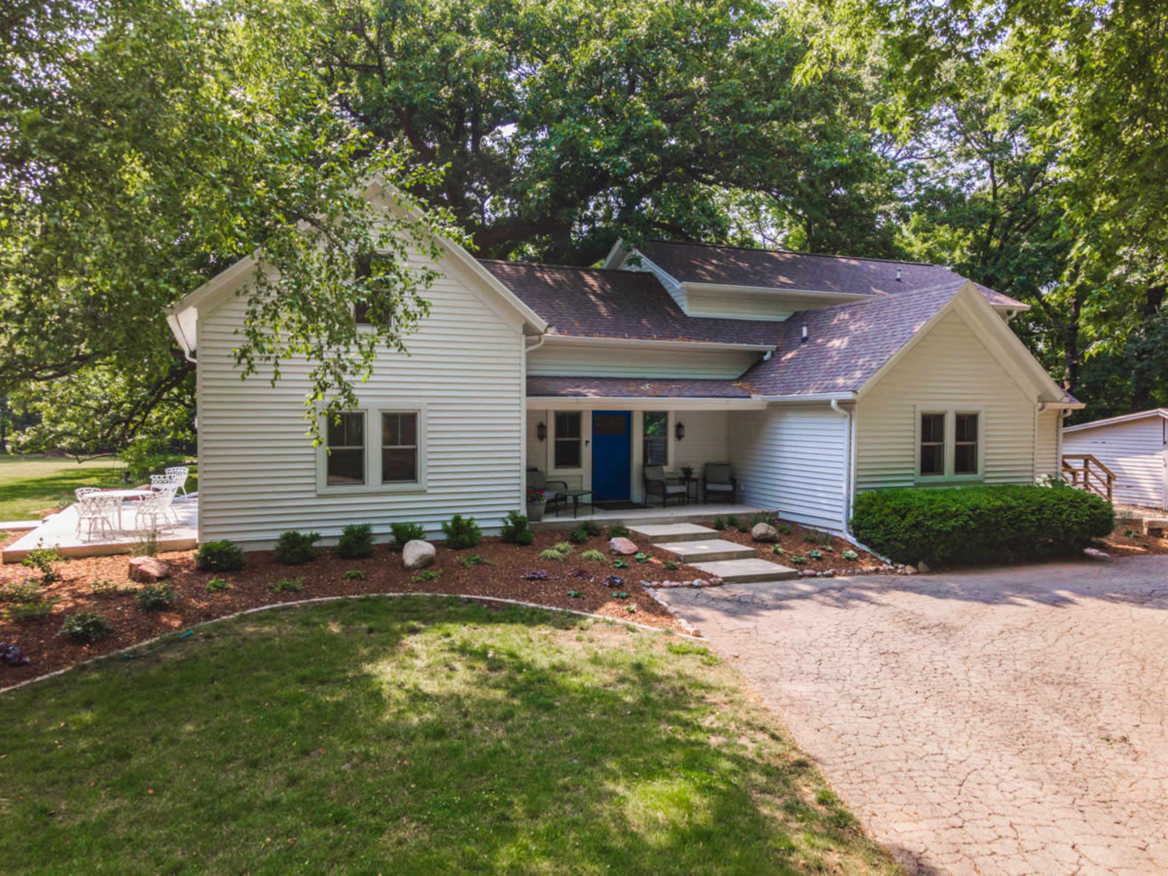 The image depicts a white house with a landscaped front yard and a blue door, surrounded by trees.