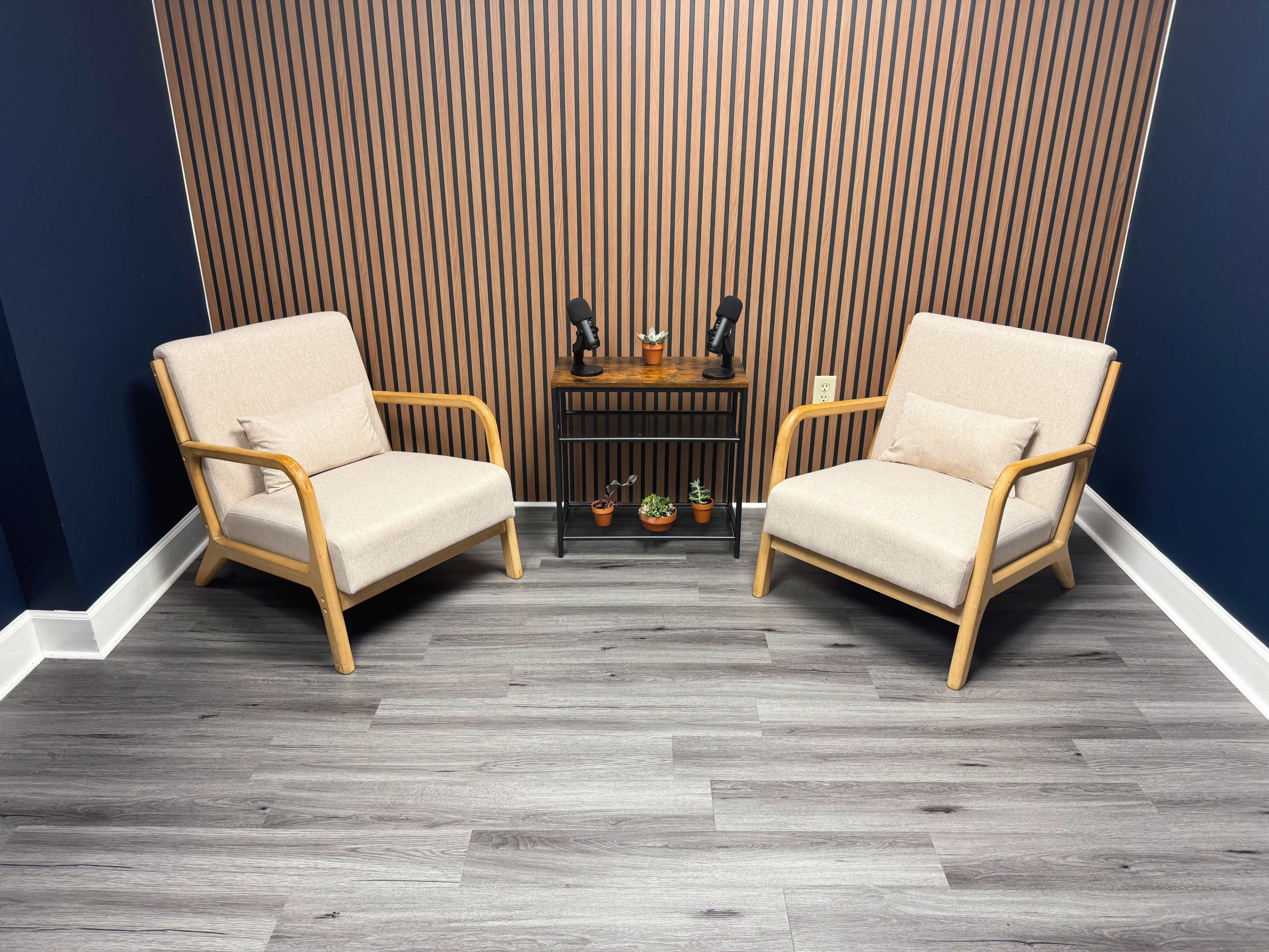 The image shows a minimalist seating arrangement with two beige armchairs and a small black table adorned with plants, set against a wood-paneled wall and gray flooring.