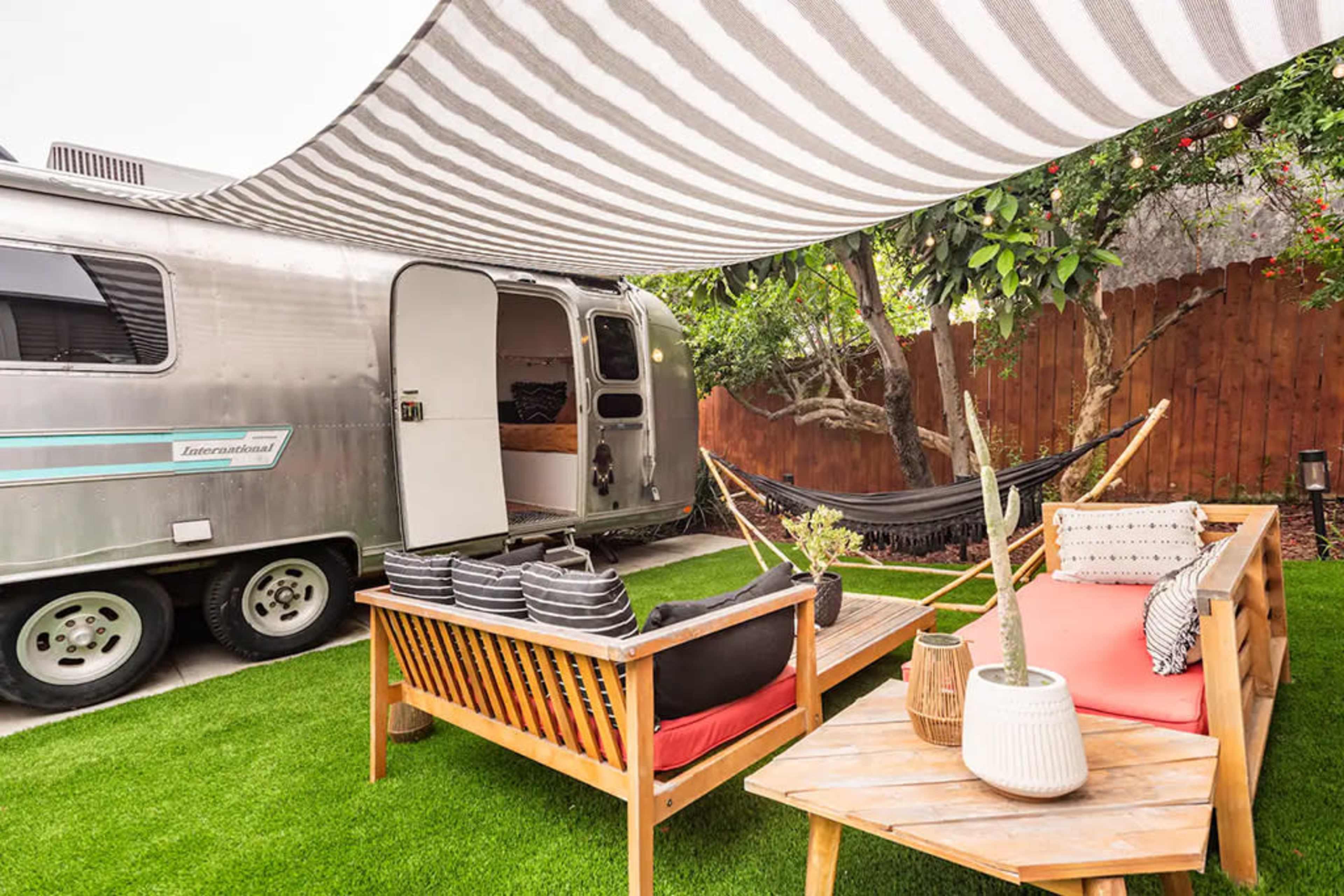 A vintage Airstream trailer is parked beside a comfortable outdoor seating area with hammocks and a striped canopy overhead.